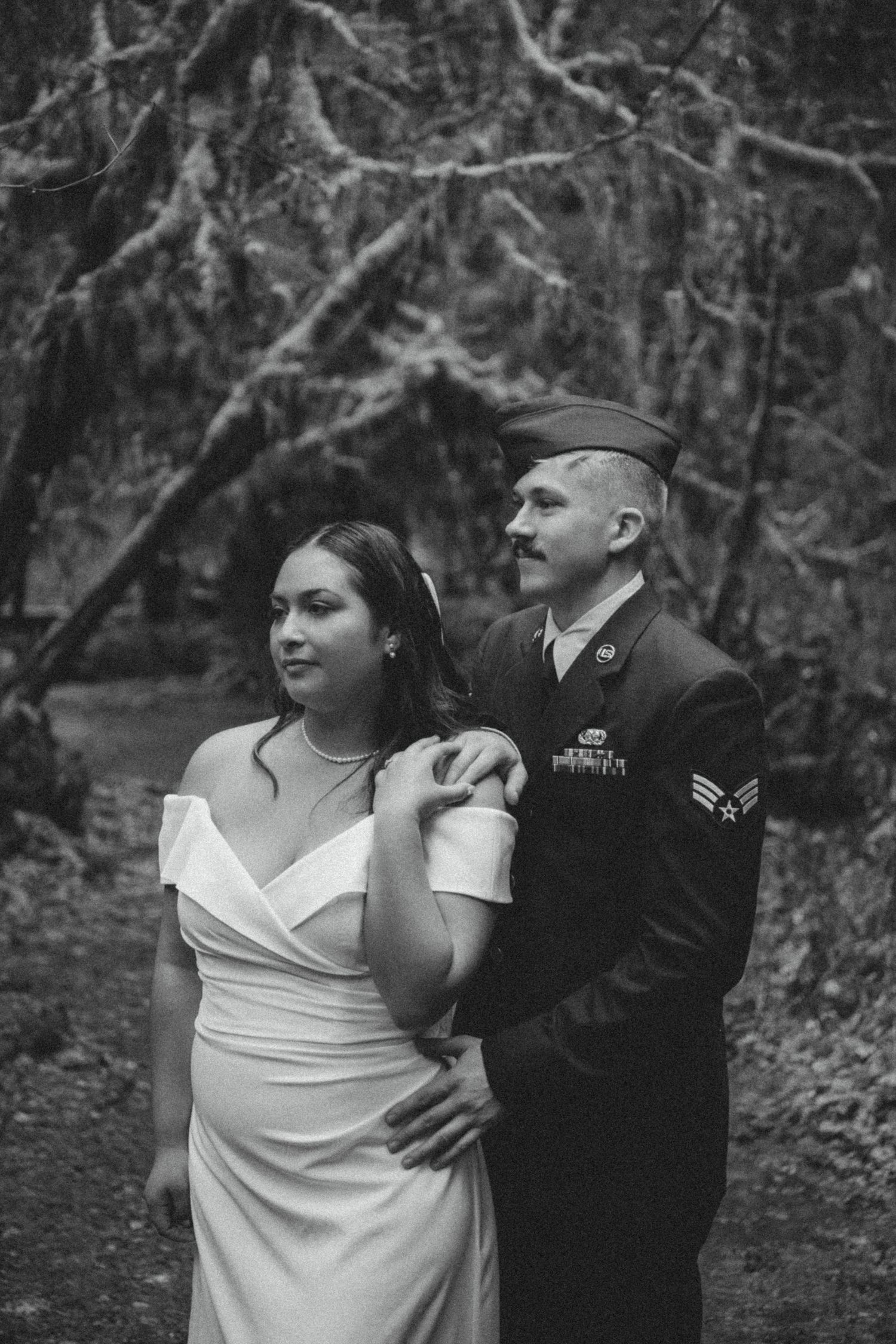 Black and white photo of a couple in a forest setting, with the man in military dress uniform and woman in white gown.