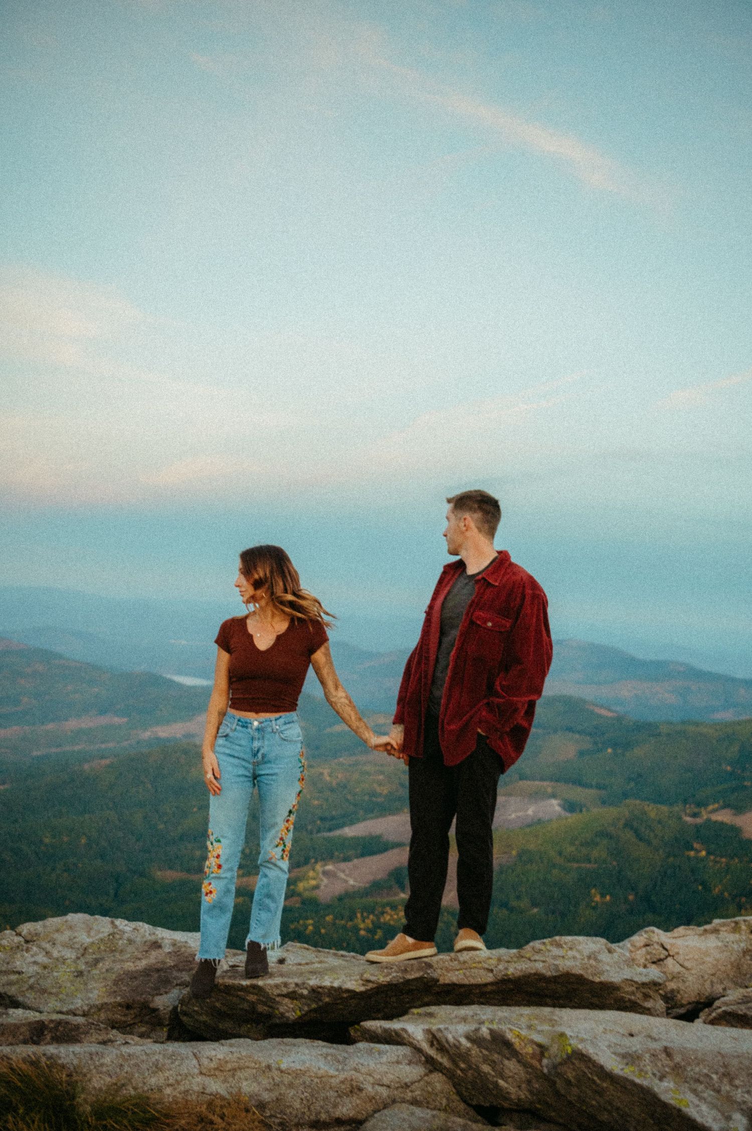 A couple stands together holding hands on a mountain summit overlooking misty hills at sunset.