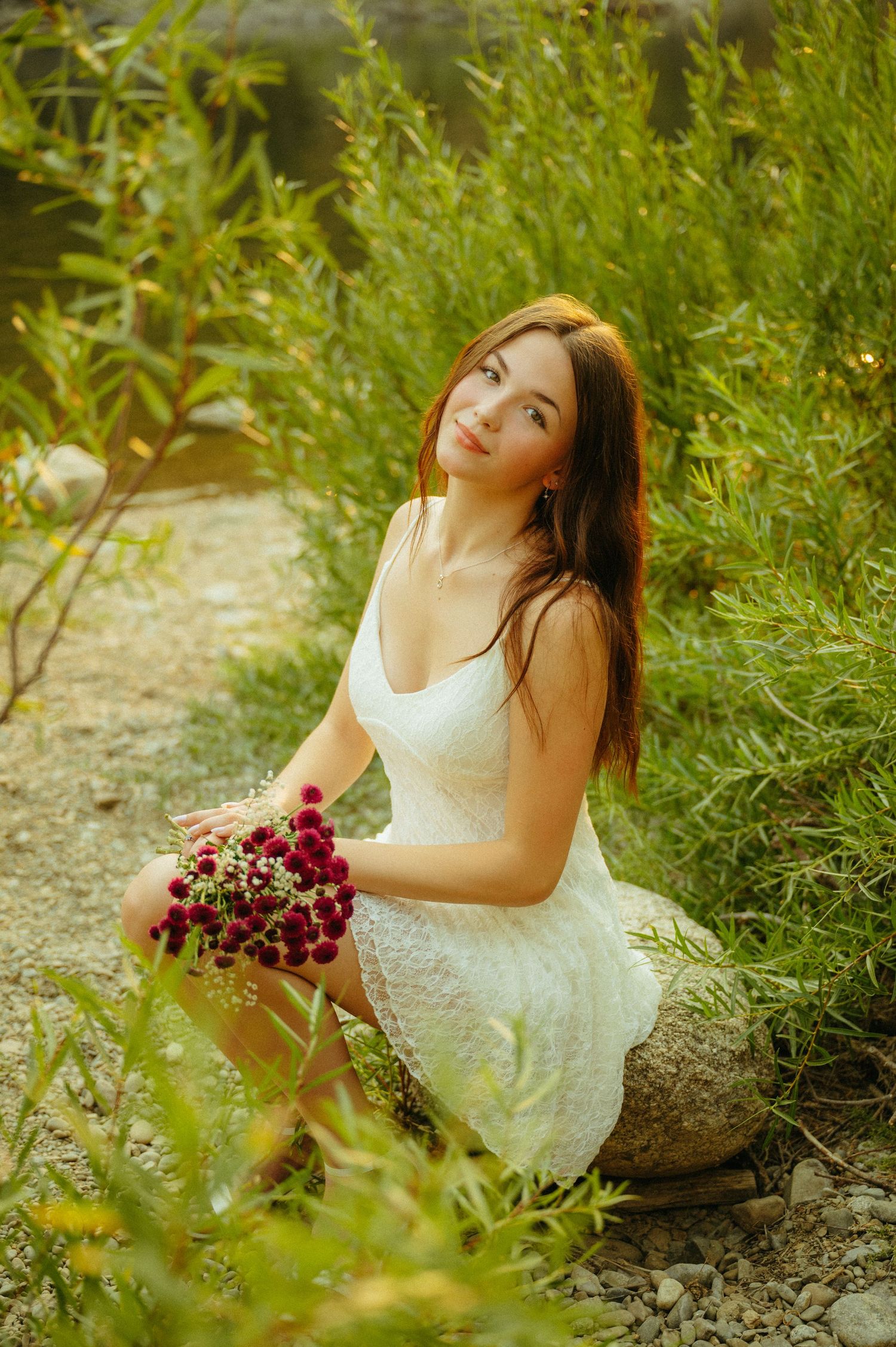 A person in a white dress sits in a garden holding a bouquet of burgundy flowers on a summer evening.