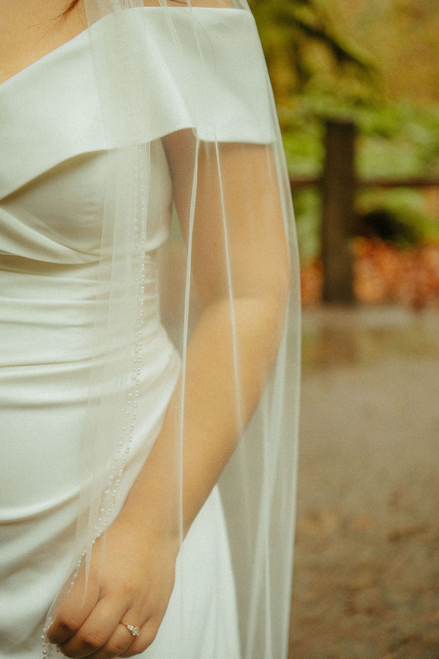 Close-up detail of a white wedding dress with flowing fabric and a delicate ring on the hand in an outdoor setting.