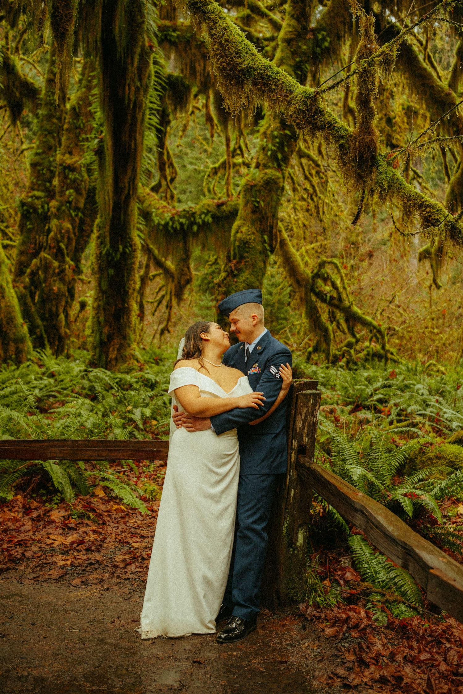 A romantic wedding couple embraces on a wooden bridge surrounded by moss-covered trees in a lush rainforest setting.