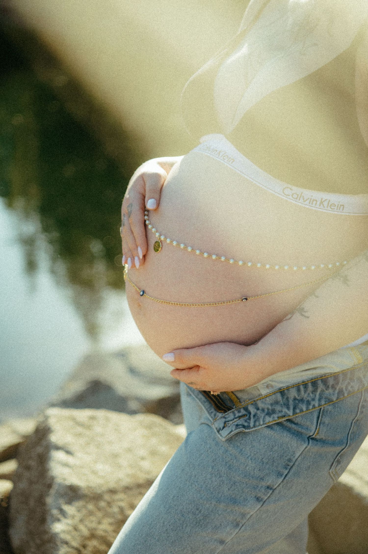 A pregnant belly adorned with jewelry is photographed against a natural outdoor setting with sunlight and rocks.