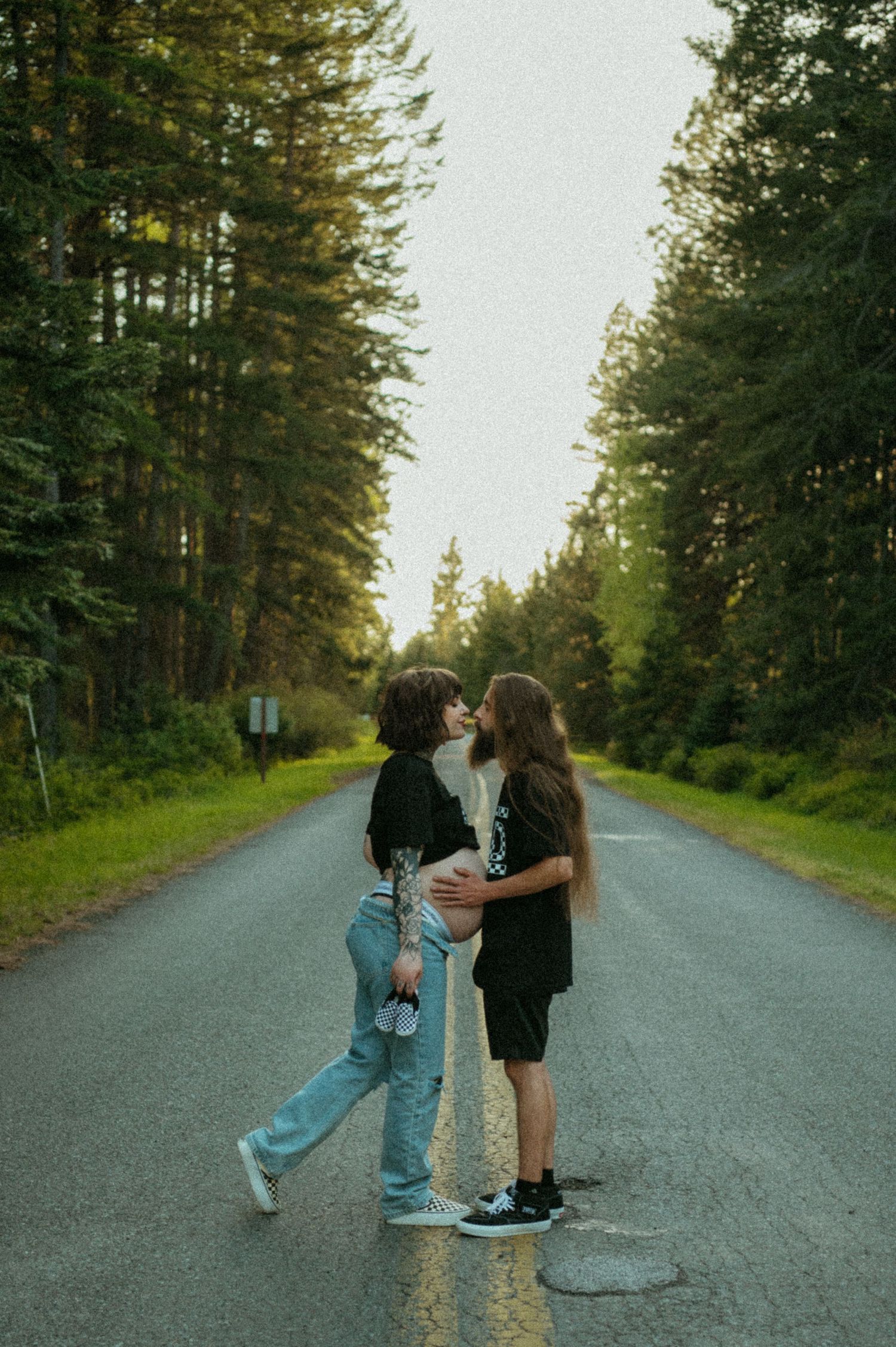 Two people embracing on a tree-lined rural road during golden hour with evergreen forest in the background.