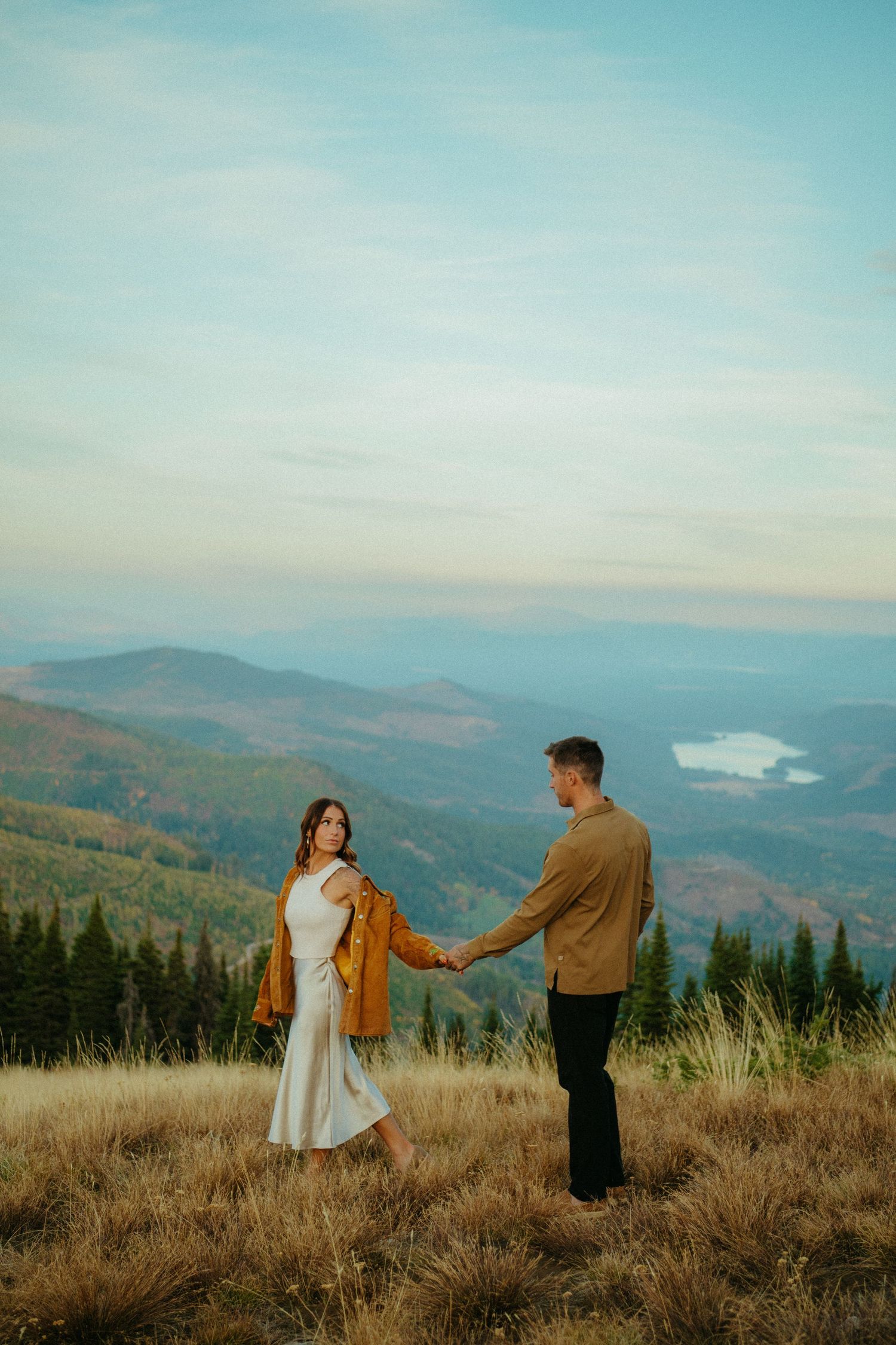 A couple holds hands on a mountaintop meadow overlooking distant hills and a lake during golden hour.