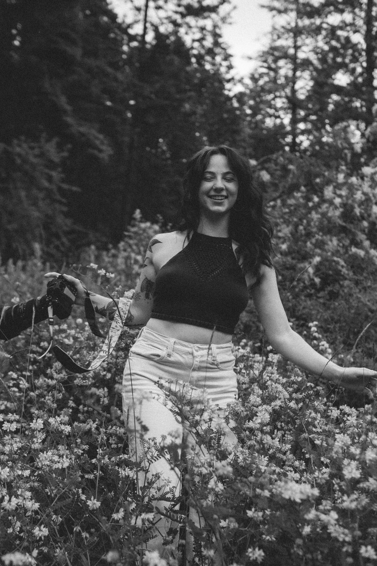 Black and white photo of someone in a crop top and high-waisted pants standing among wildflowers in a forest setting.