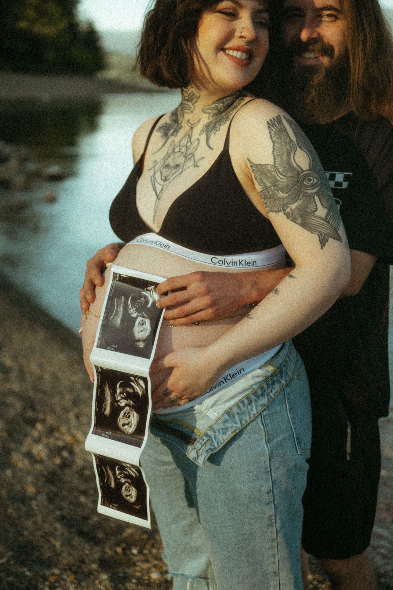 A couple standing by a lake holds ultrasound photos while showing off tattoos and a baby bump in casual attire.