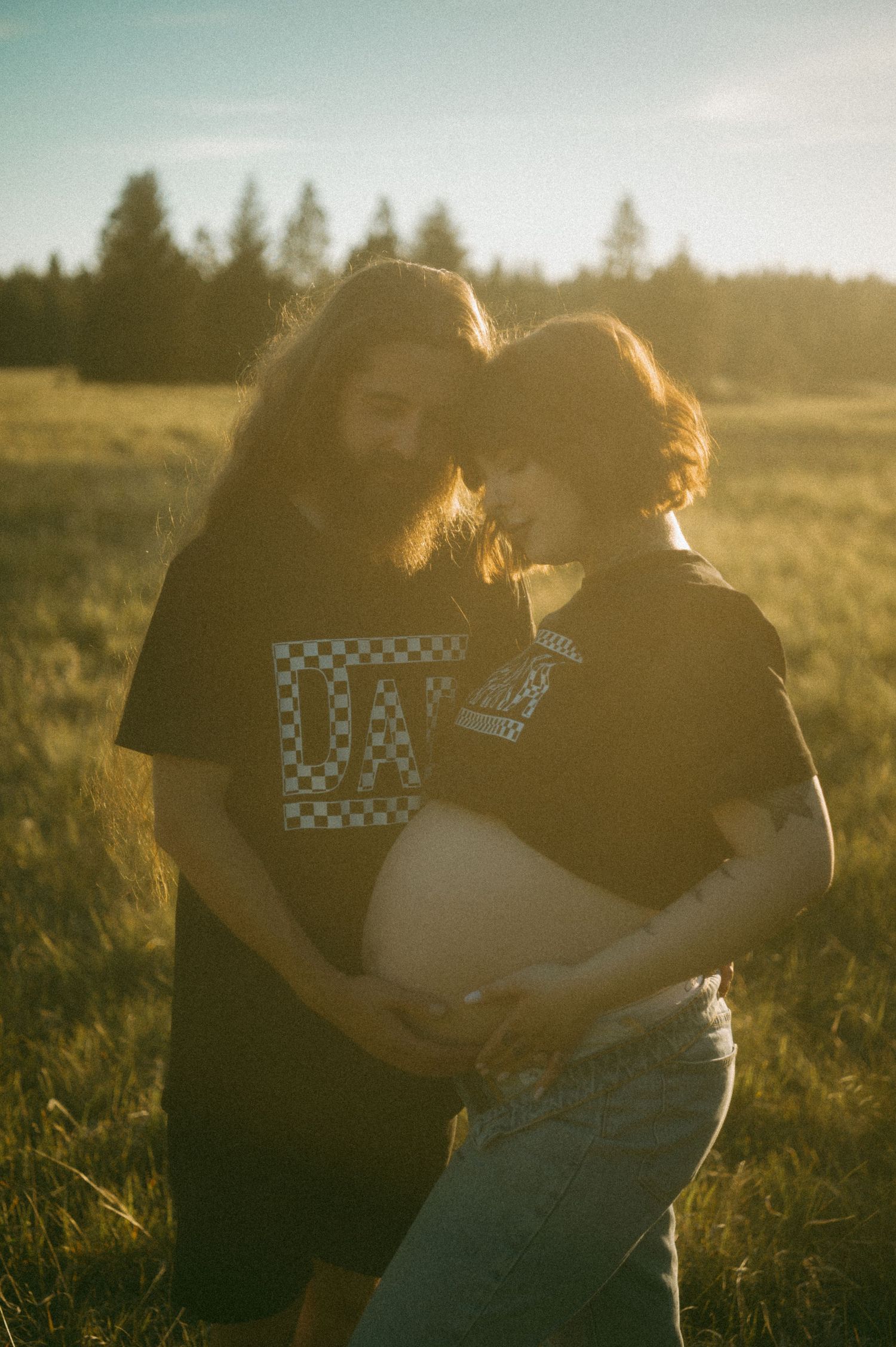 Two people embracing outdoors in a grassy field at sunset with backlit hair and casual clothing.