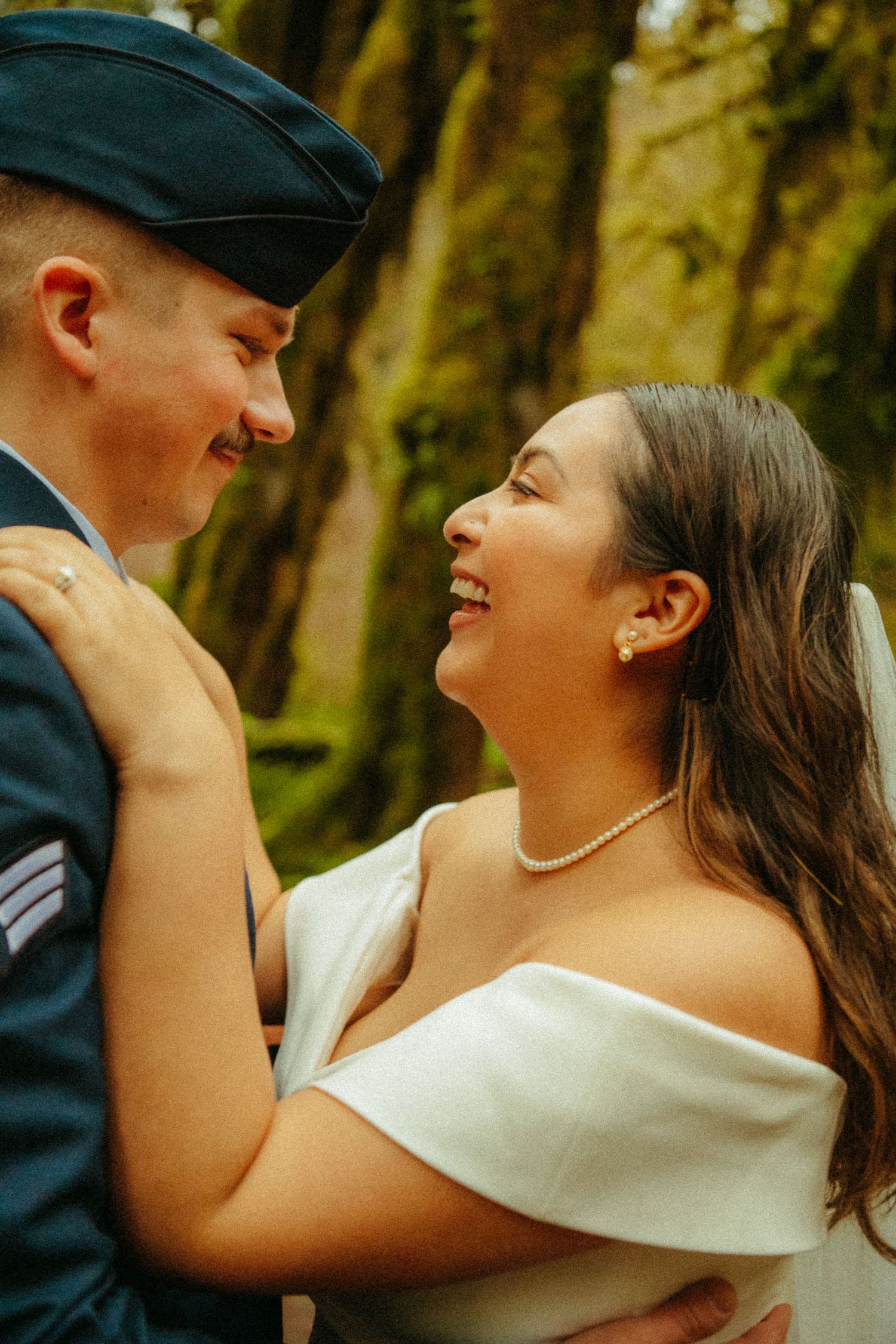 A joyful wedding couple shares a tender moment laughing together in a wooded outdoor setting.