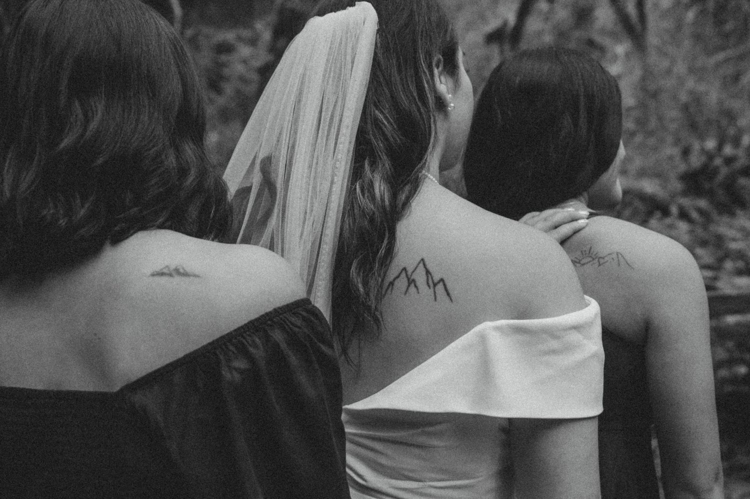 Black and white image showing three shoulders of wedding guests with dress details and a flowing veil in view.