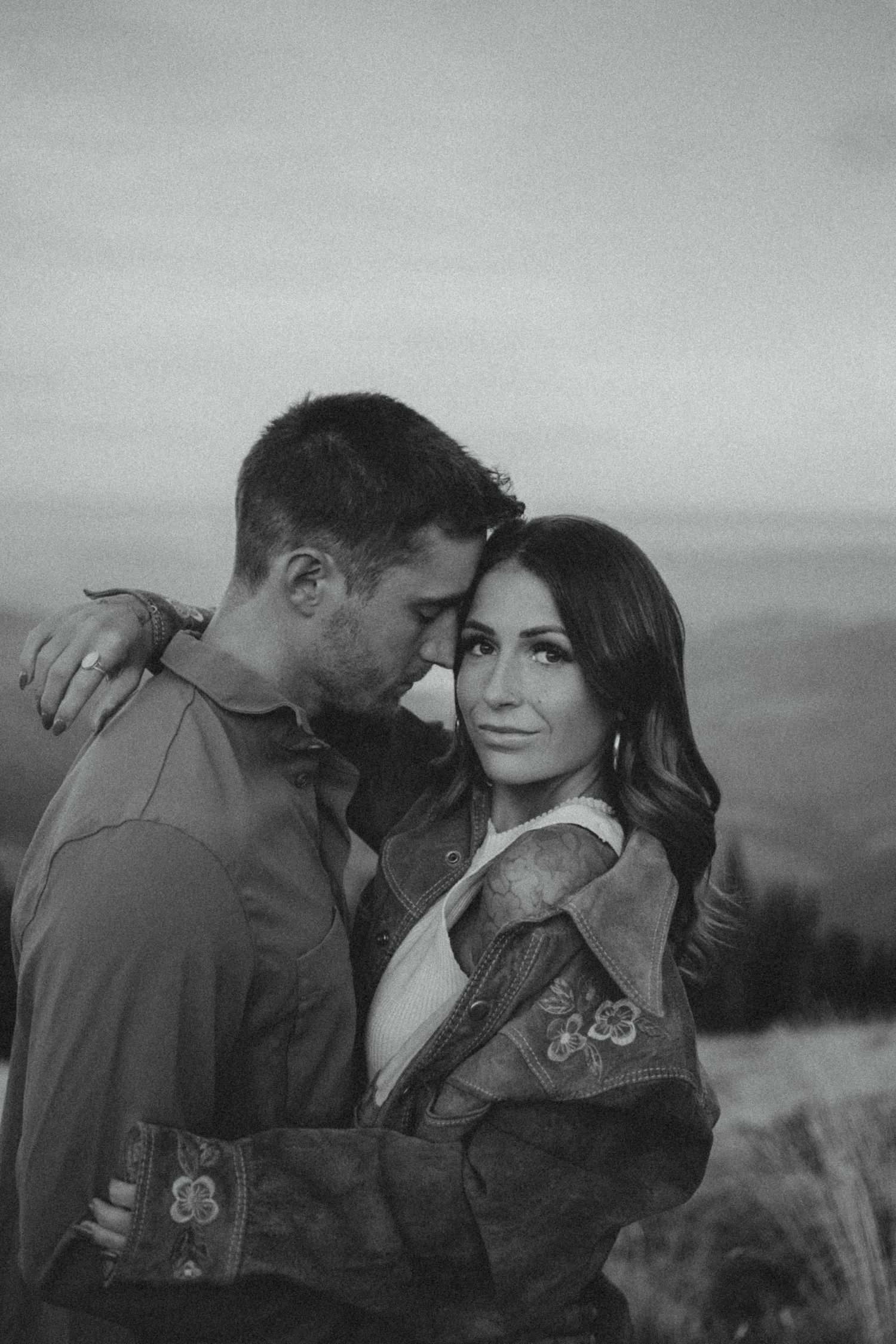 Black and white romantic portrait of a couple embracing outdoors with a scenic mountain view in the background.