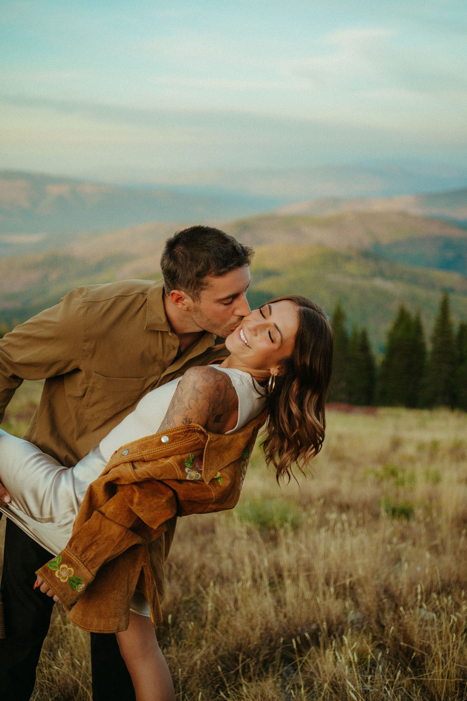 A romantic couple embraces and shares a tender moment against a mountainous backdrop at sunset.