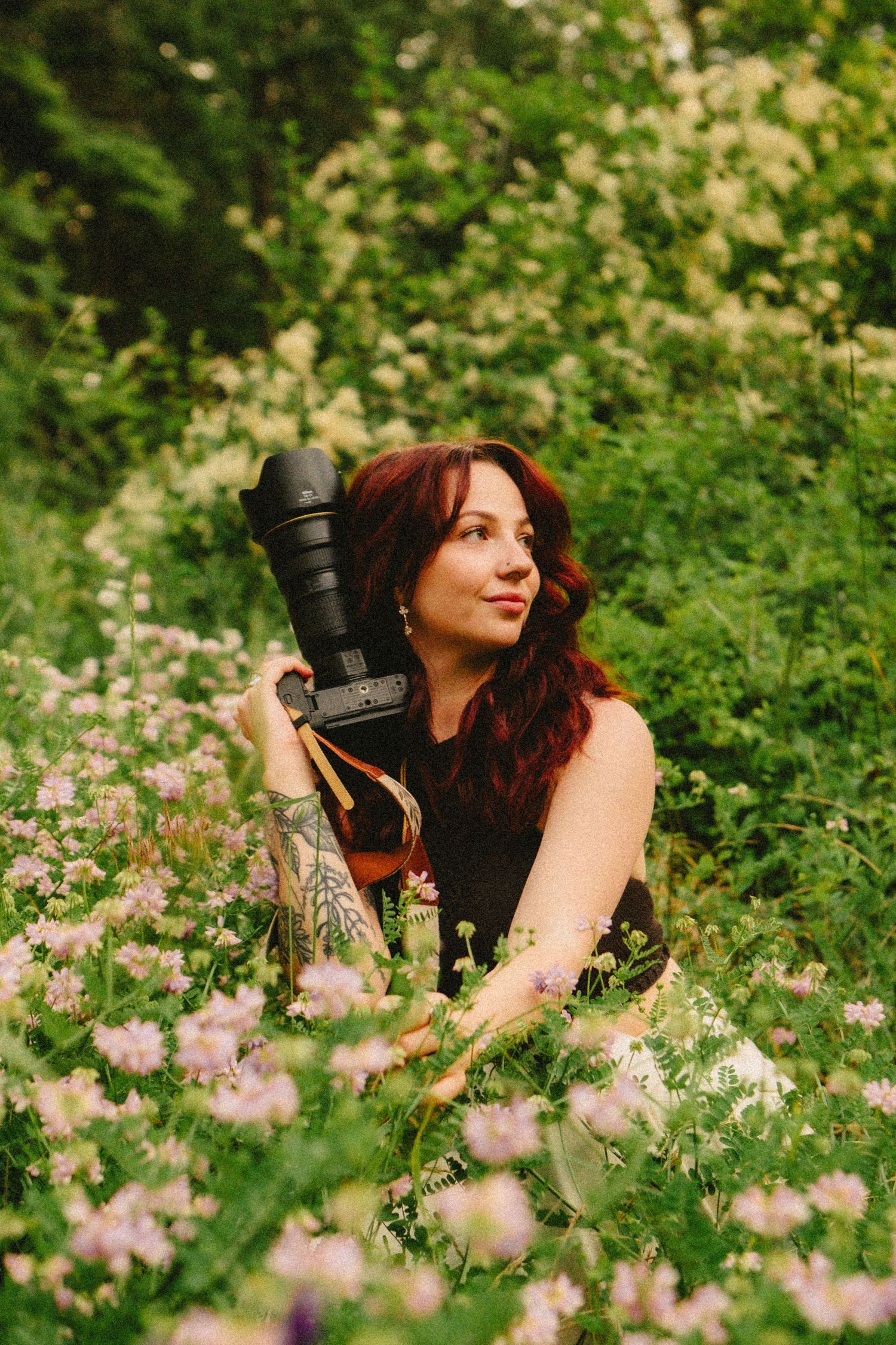 Photographer with long red hair holds professional camera while sitting among pink wildflowers in a lush garden setting.