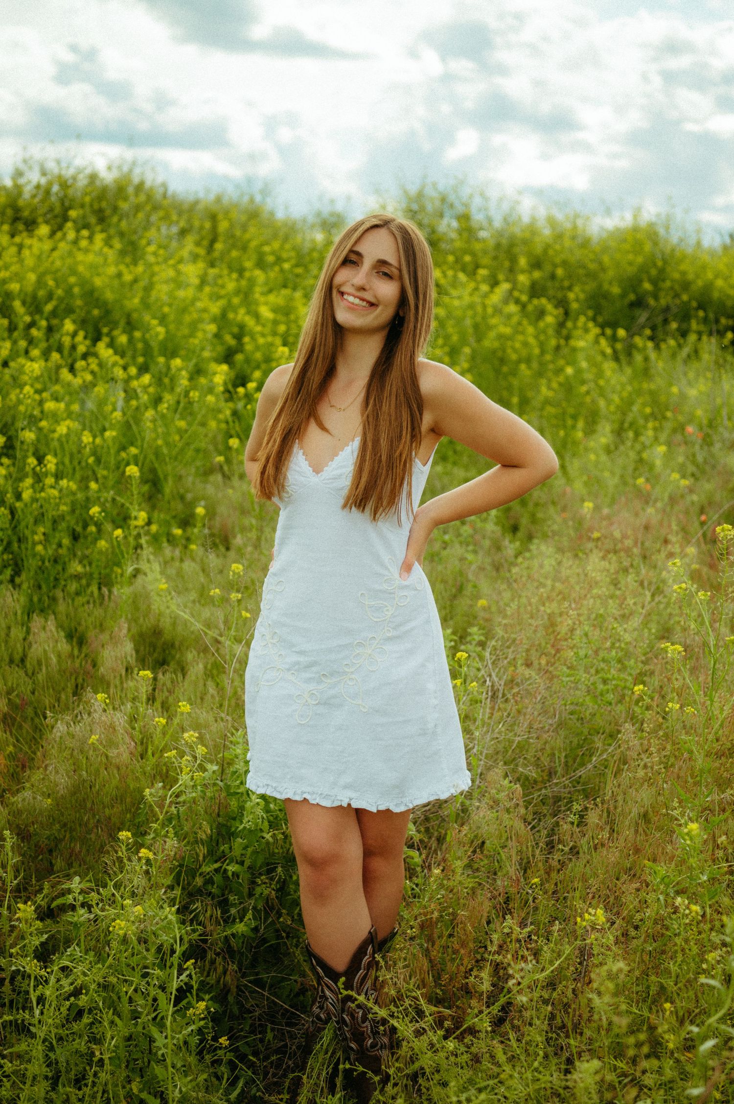 A person in a white lace dress poses playfully in a blooming yellow canola field on a cloudy summer day.