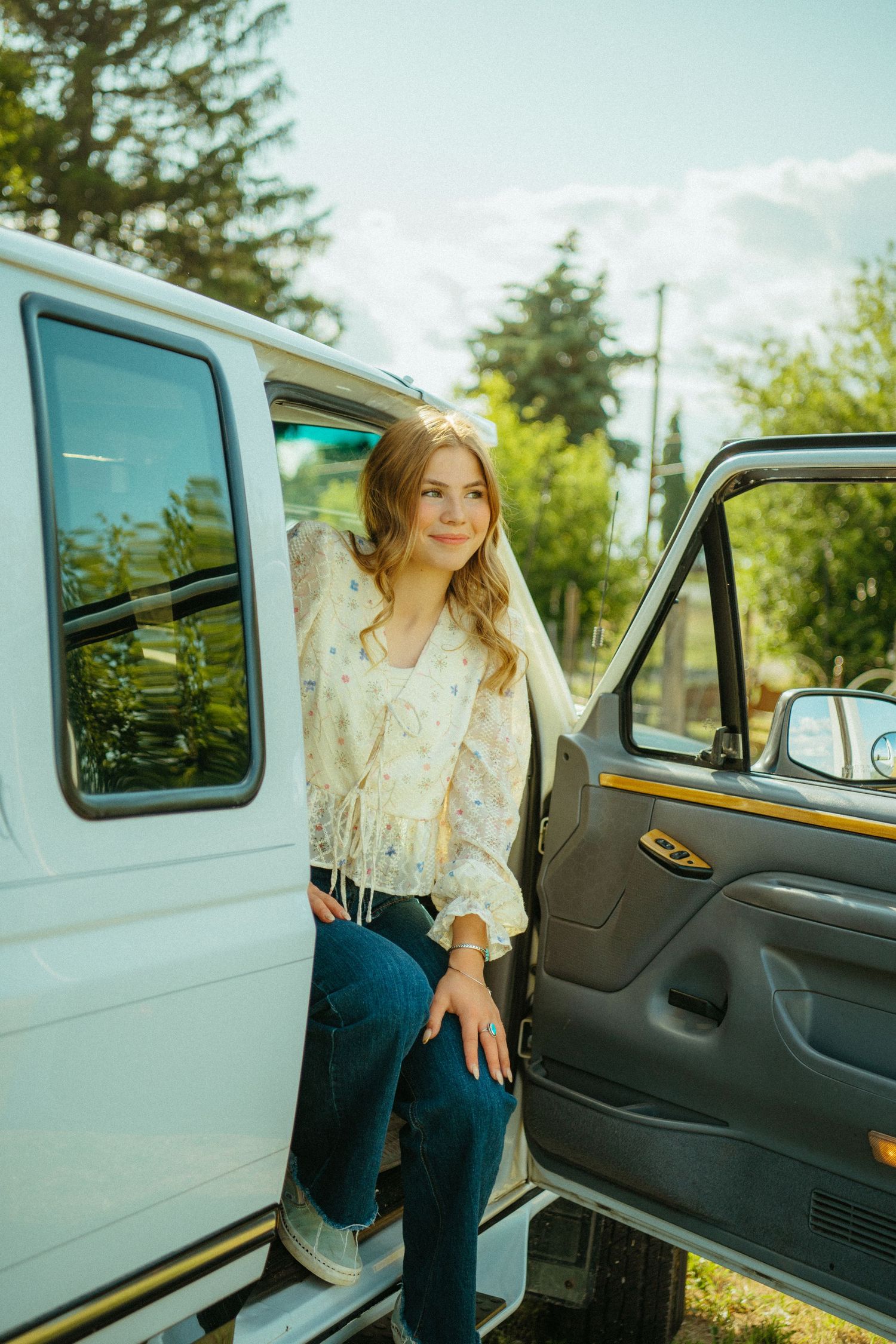 A person in a white blouse and jeans sits in a vintage car with open door on a sunny day.