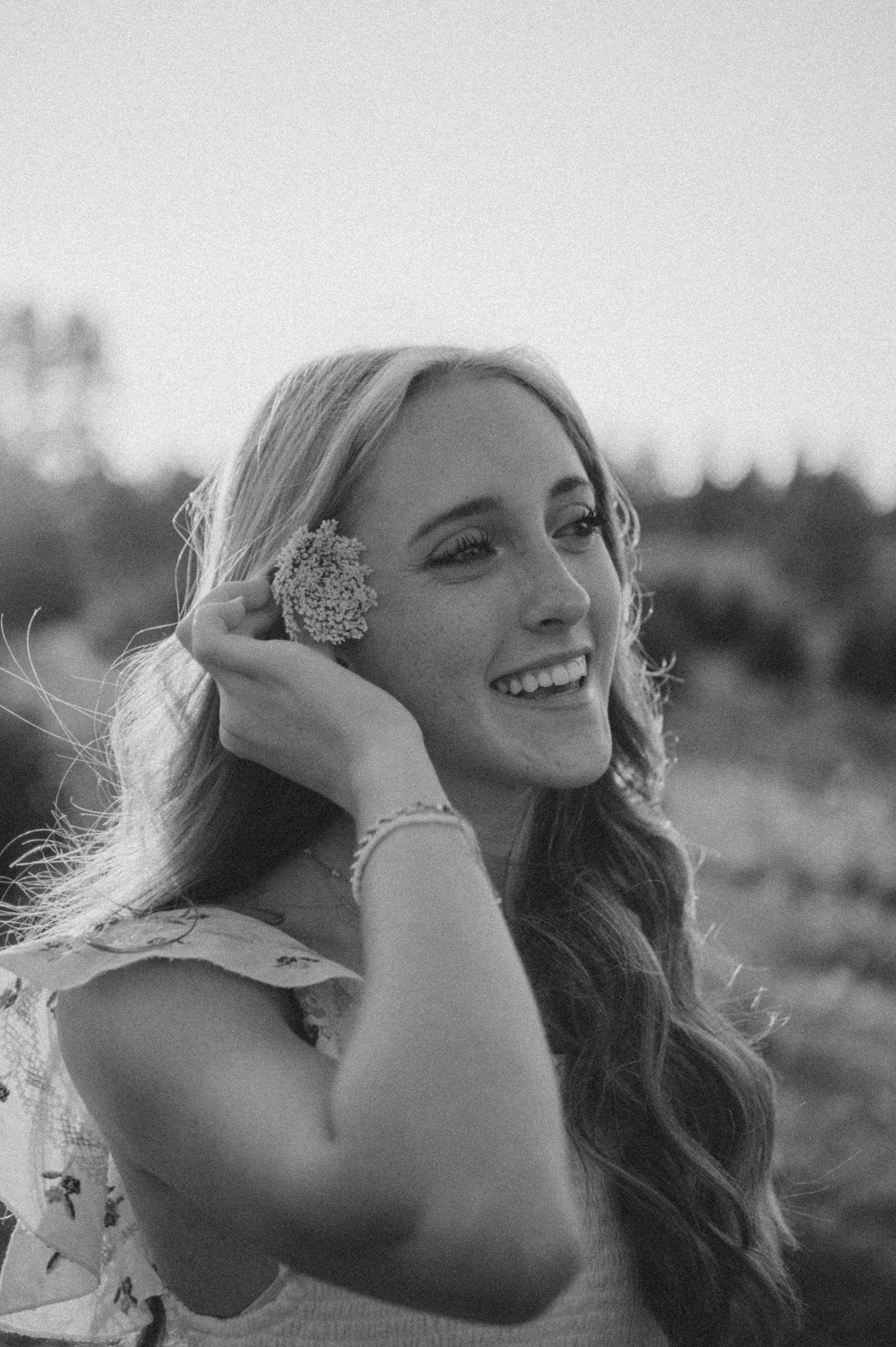 A black and white portrait of a smiling woman in a dress with long wavy hair and floral accessories.