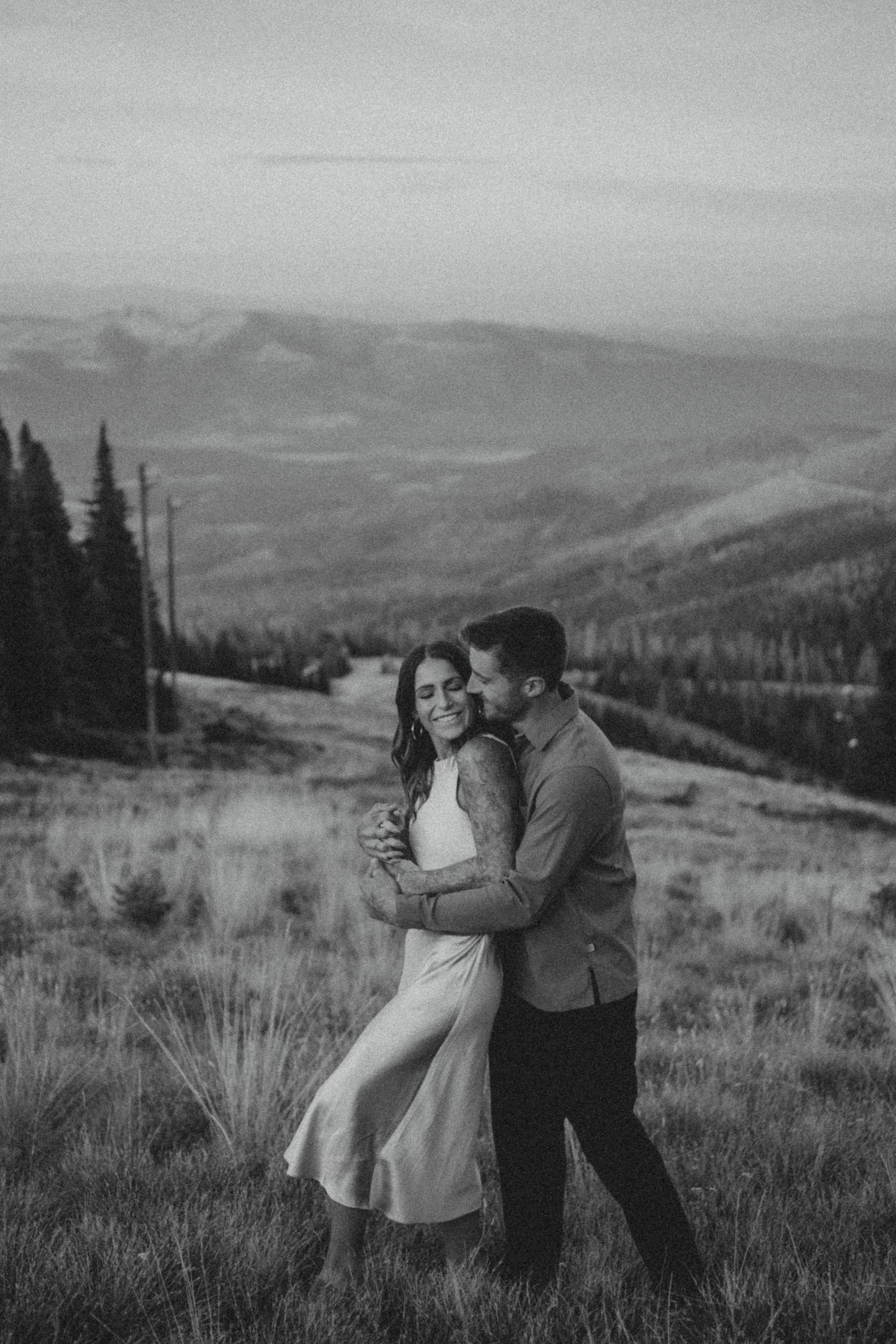 A romantic black and white mountain scene with a couple embracing on a hillside against a backdrop of evergreen trees.