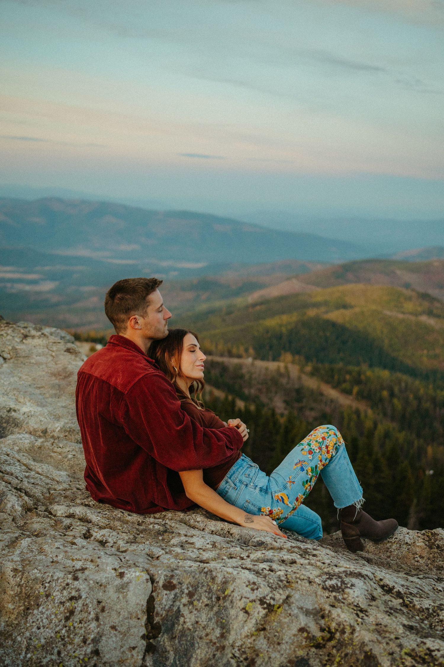 Couple relaxing on mountain cliff edge during sunset overlooking scenic mountainous valley with forest below.