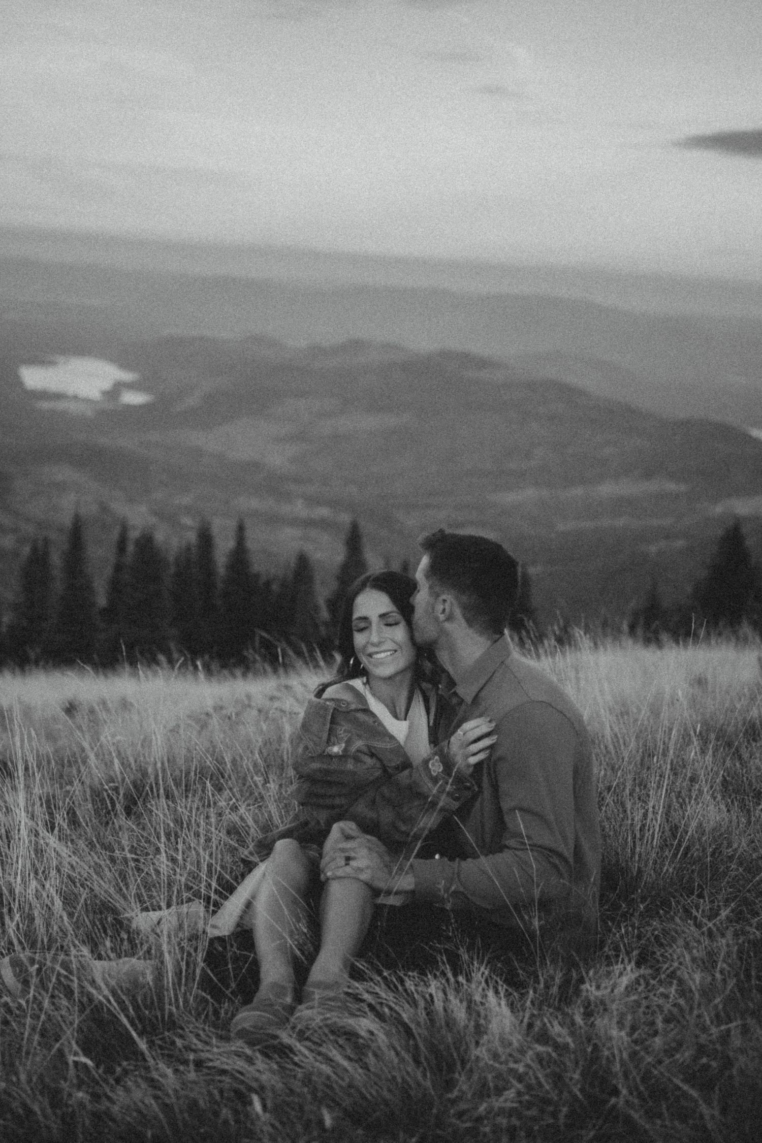 A couple sits together on a hillside with mountains and evergreen trees in the distance at dusk, captured in black and white.