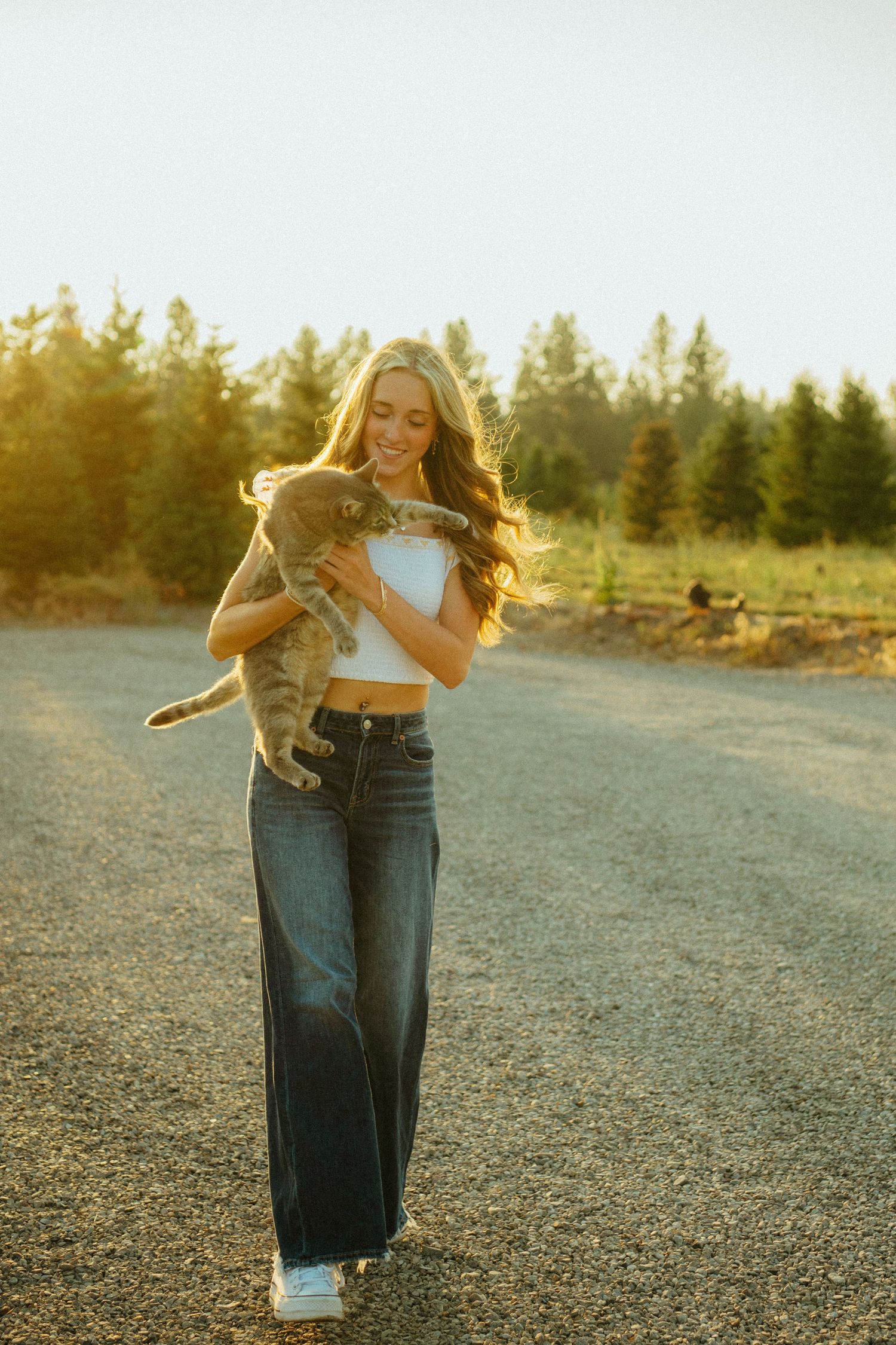 Stylish person wearing crop top and wide-leg jeans holds small dog at sunset with evergreen trees in background.