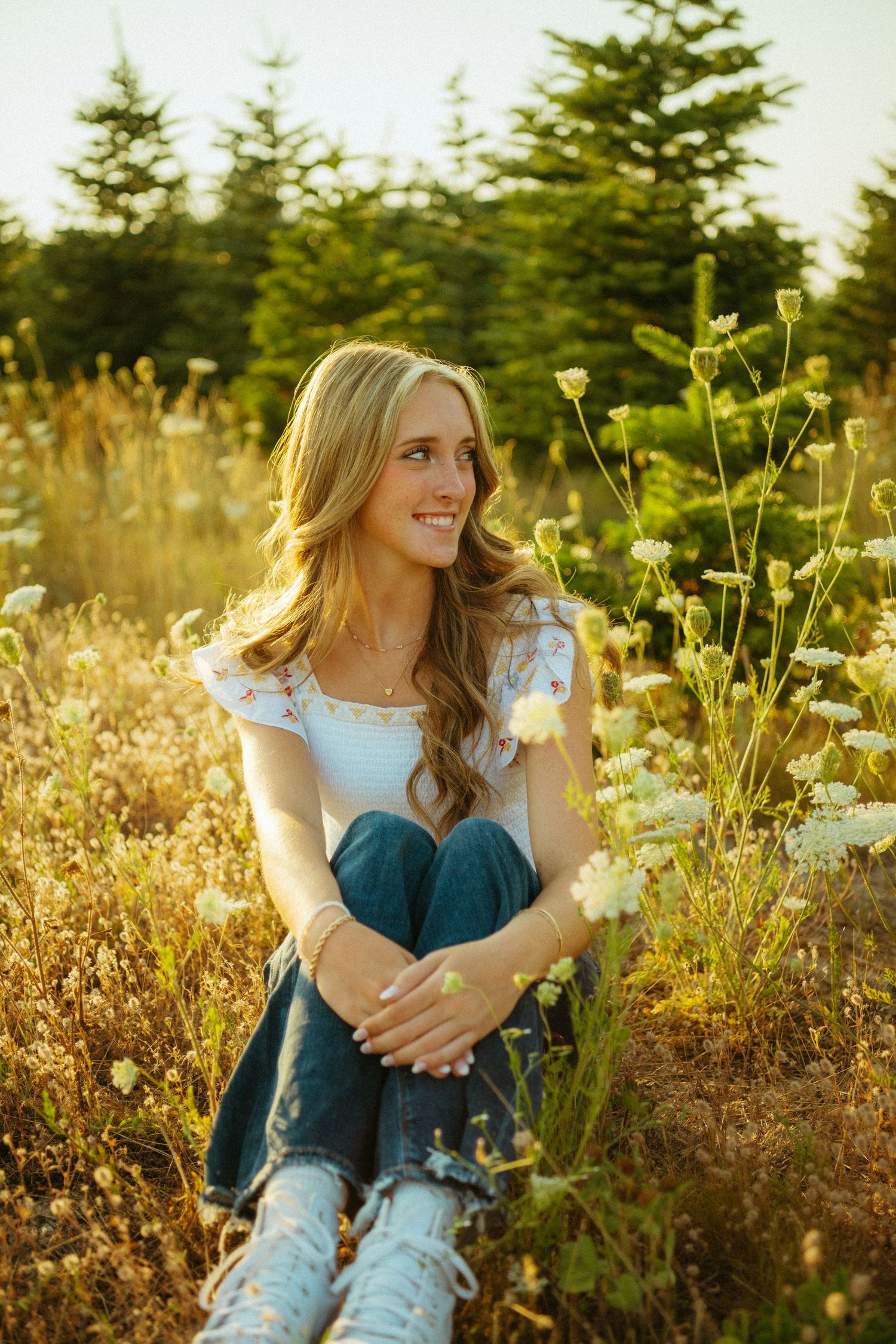 A person sits in a golden field at sunset surrounded by wildflowers and evergreen trees in the background.