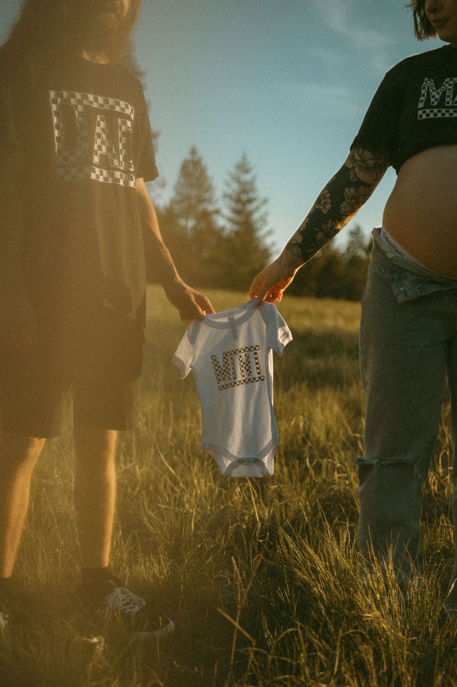 Two people hold a small white baby onesie in a sunlit field with pine trees in the background at golden hour.