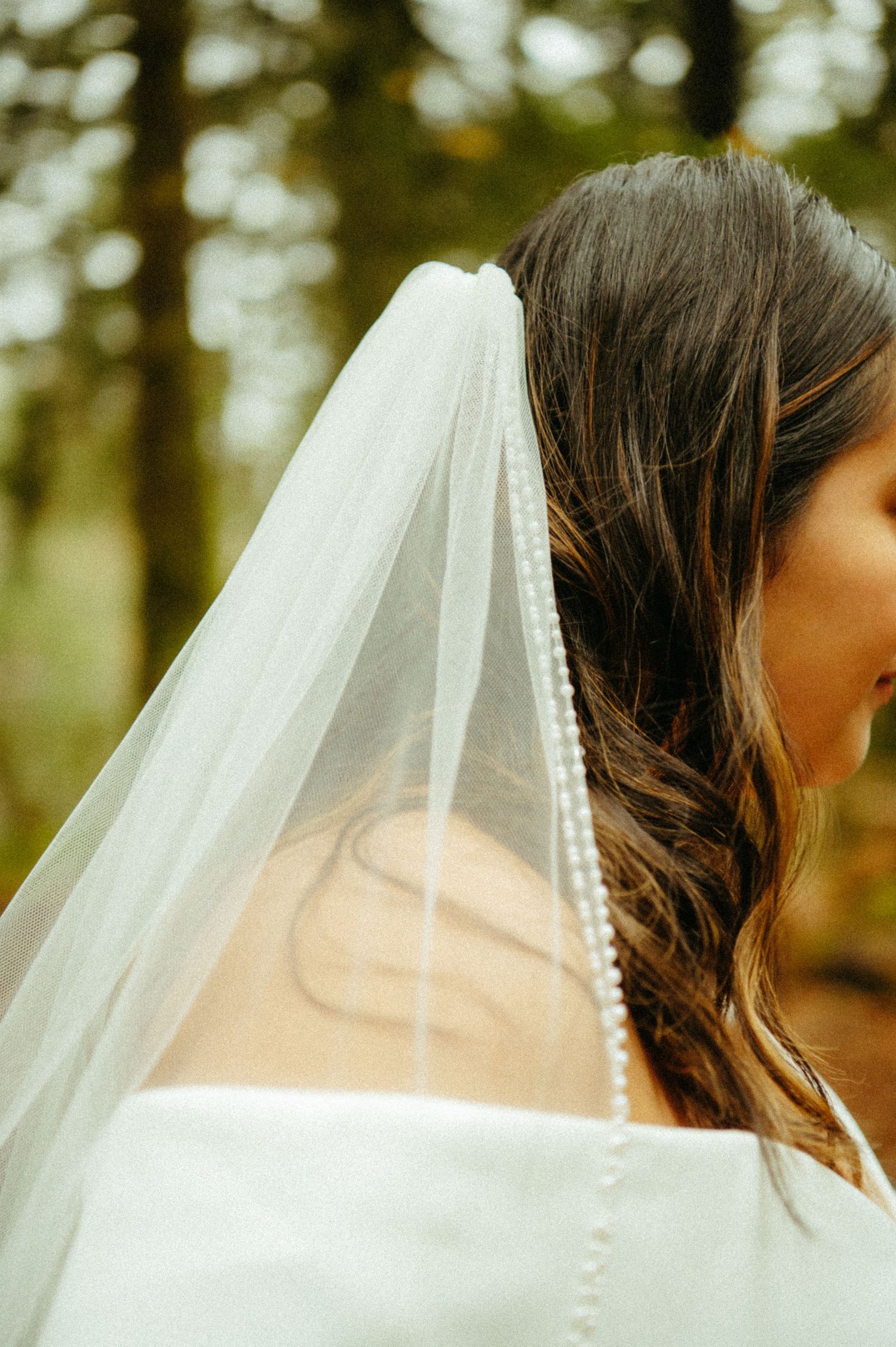 Elegant bridal veil with pearl trim drapes over dark wavy hair against a dreamy forest backdrop.