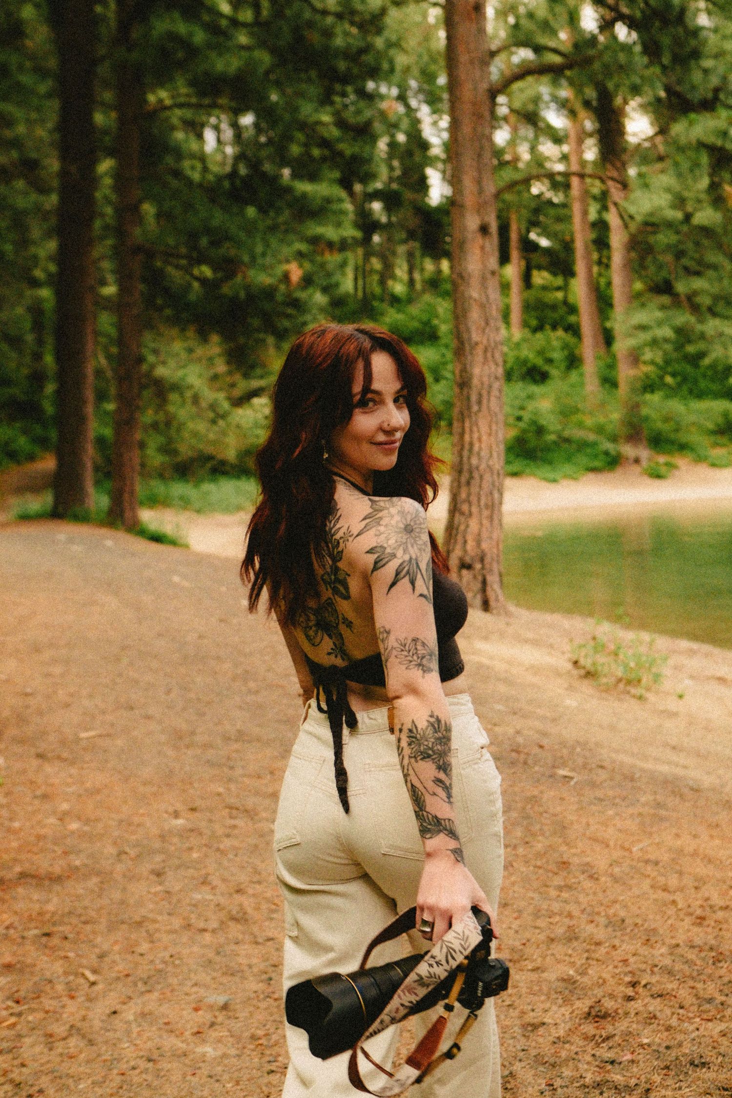 Woman with tattoos and roller skates stands on dirt path by lake surrounded by tall pine trees.