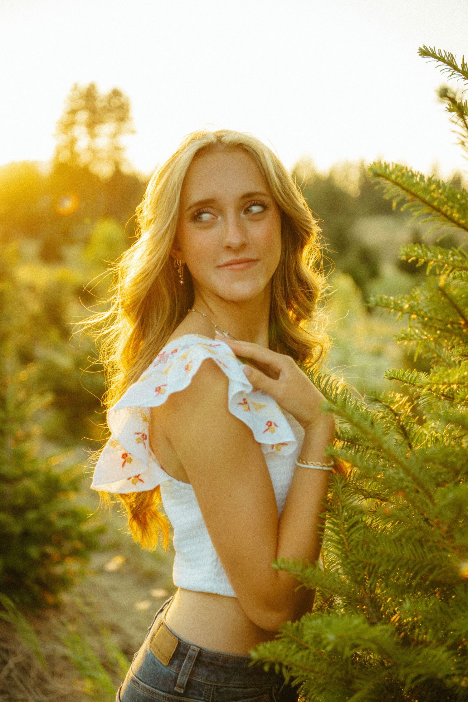 Golden sunset light illuminates portrait of young woman in white crop top by evergreen trees.