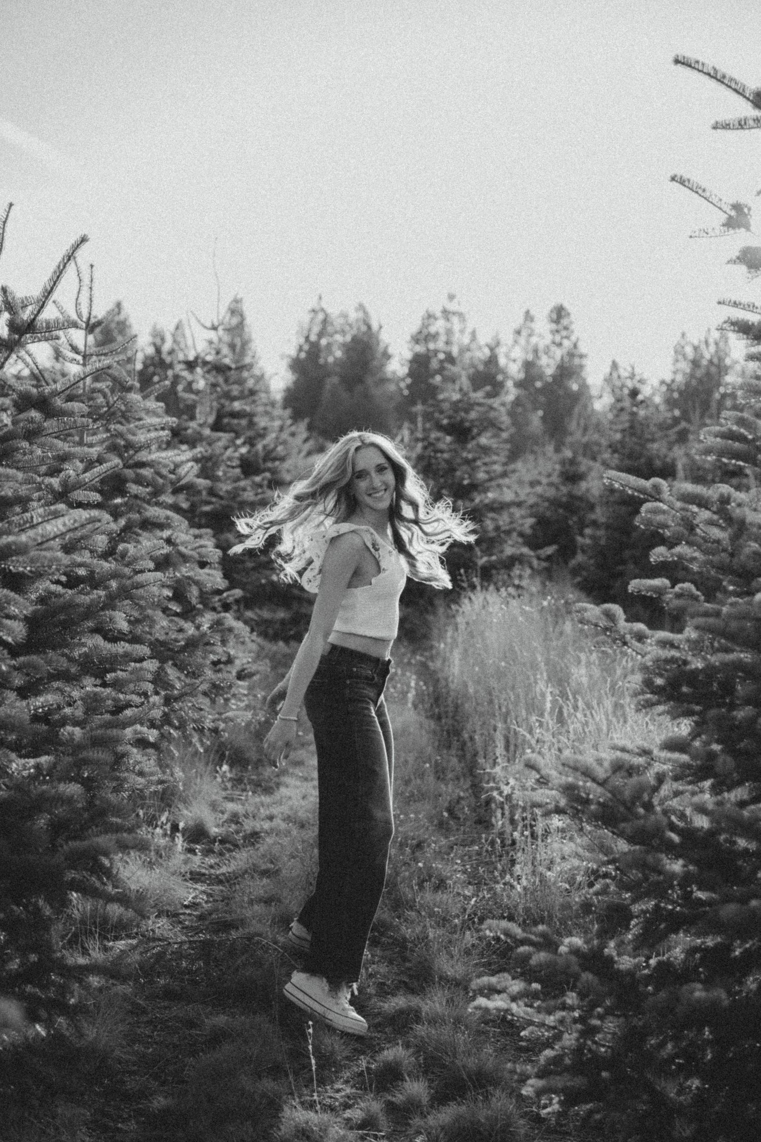 A black and white photo of someone jumping joyfully between evergreen trees at a Christmas tree farm.