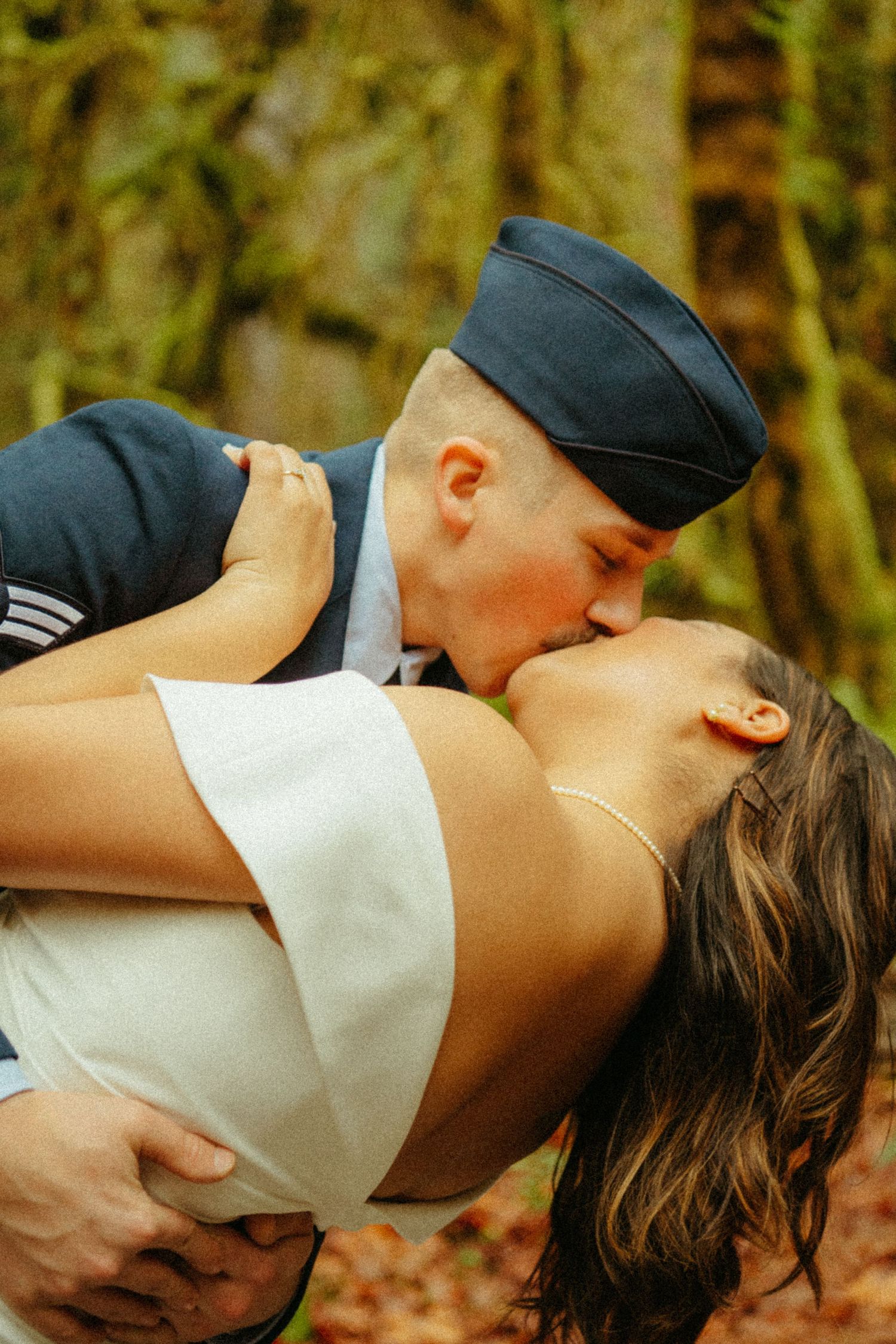 A romantic outdoor portrait shows a military servicemember in uniform kissing a woman in a white dress.