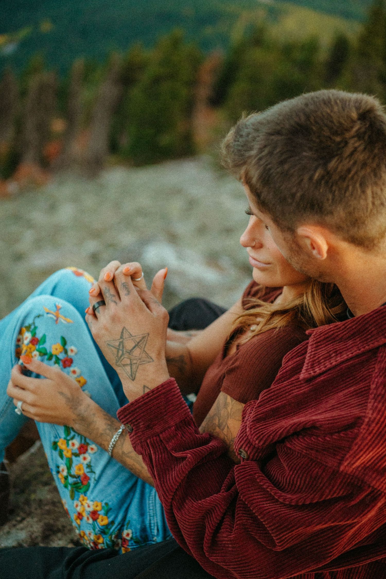 Two people share an intimate moment outdoors wearing a burgundy sweater and floral patterned pants at sunset.