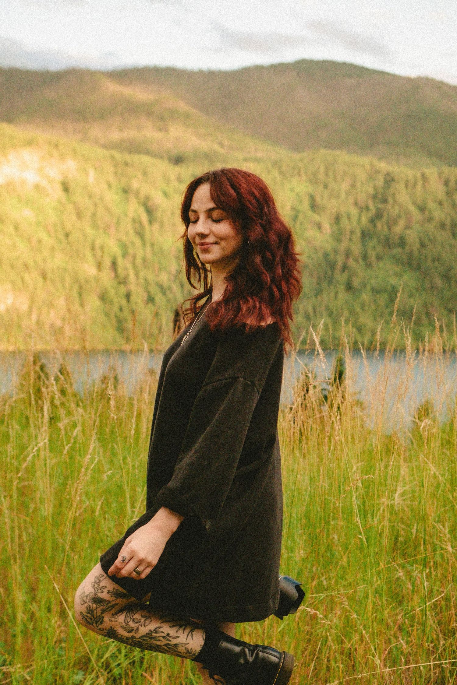 Someone in a black dress stands in tall grass by a lake with mountains in the background during golden hour.