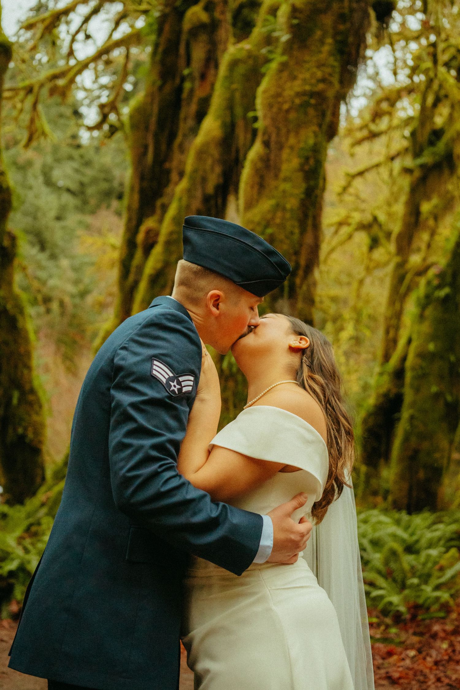 A romantic wedding couple shares a kiss in a mossy forest with lush green trees and natural lighting.