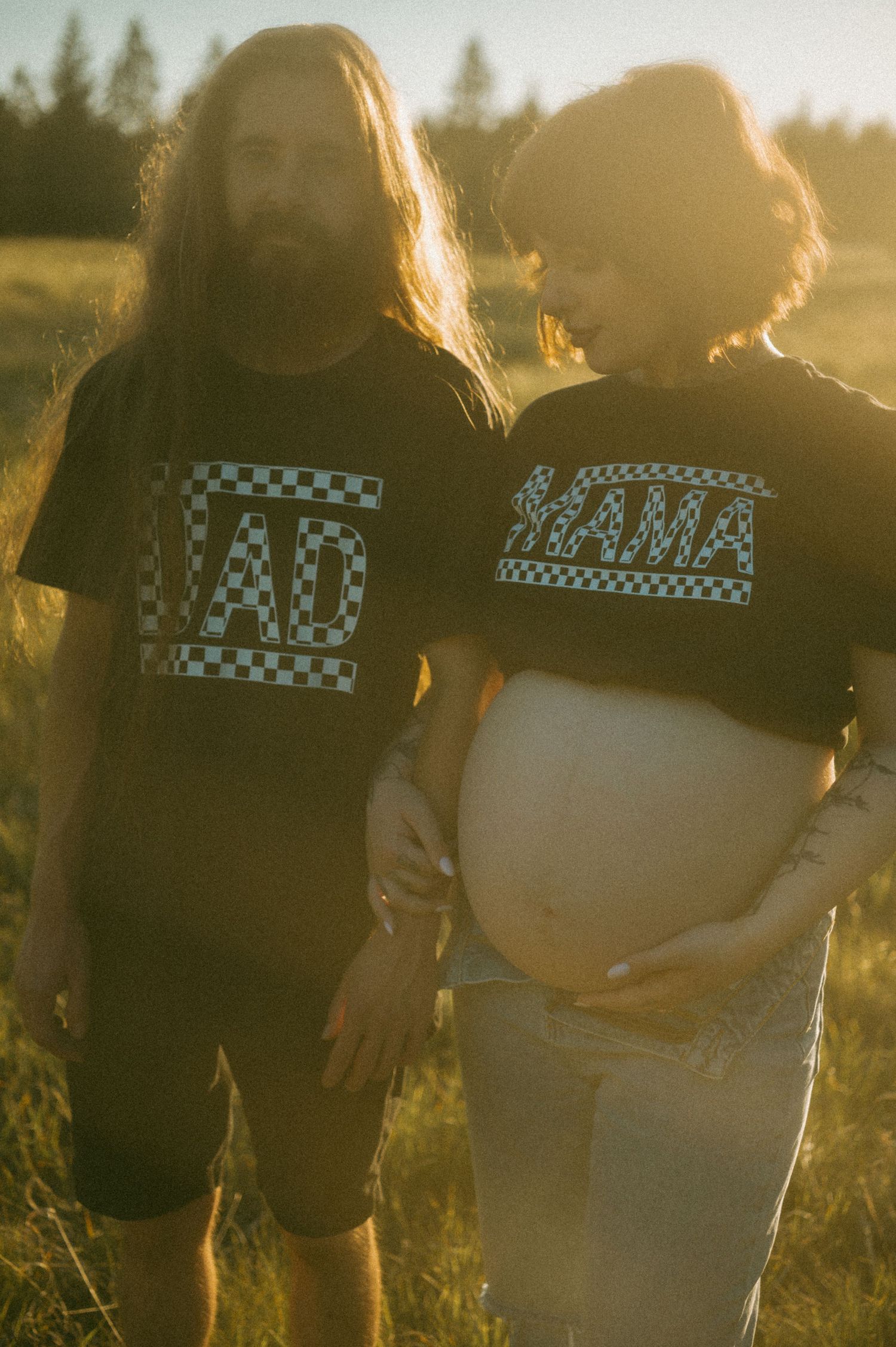 A couple wearing matching checkered text t-shirts poses together in a sunlit field at golden hour.