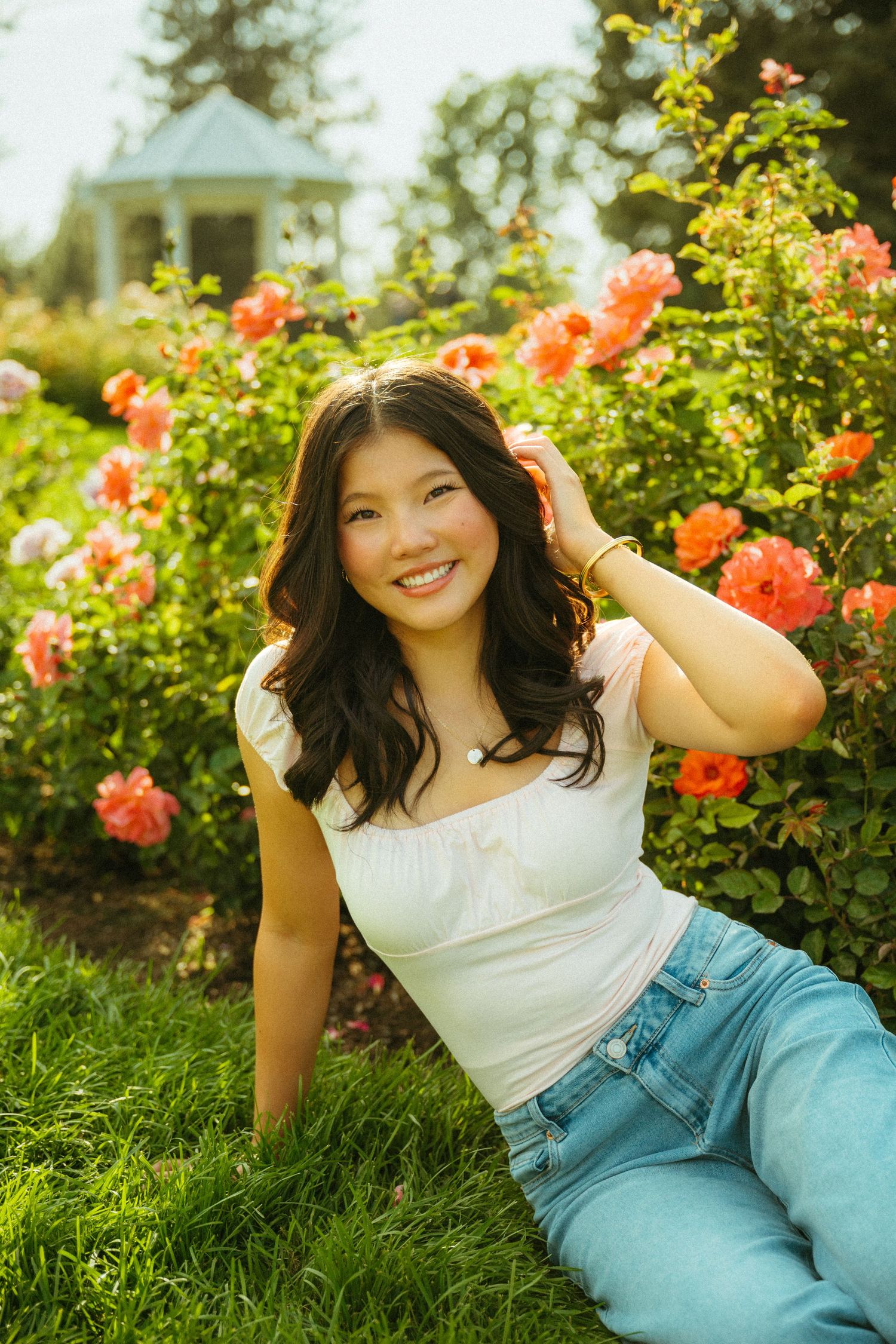 A person in a white top and blue jeans poses playfully in a garden with blooming orange roses and greenery.