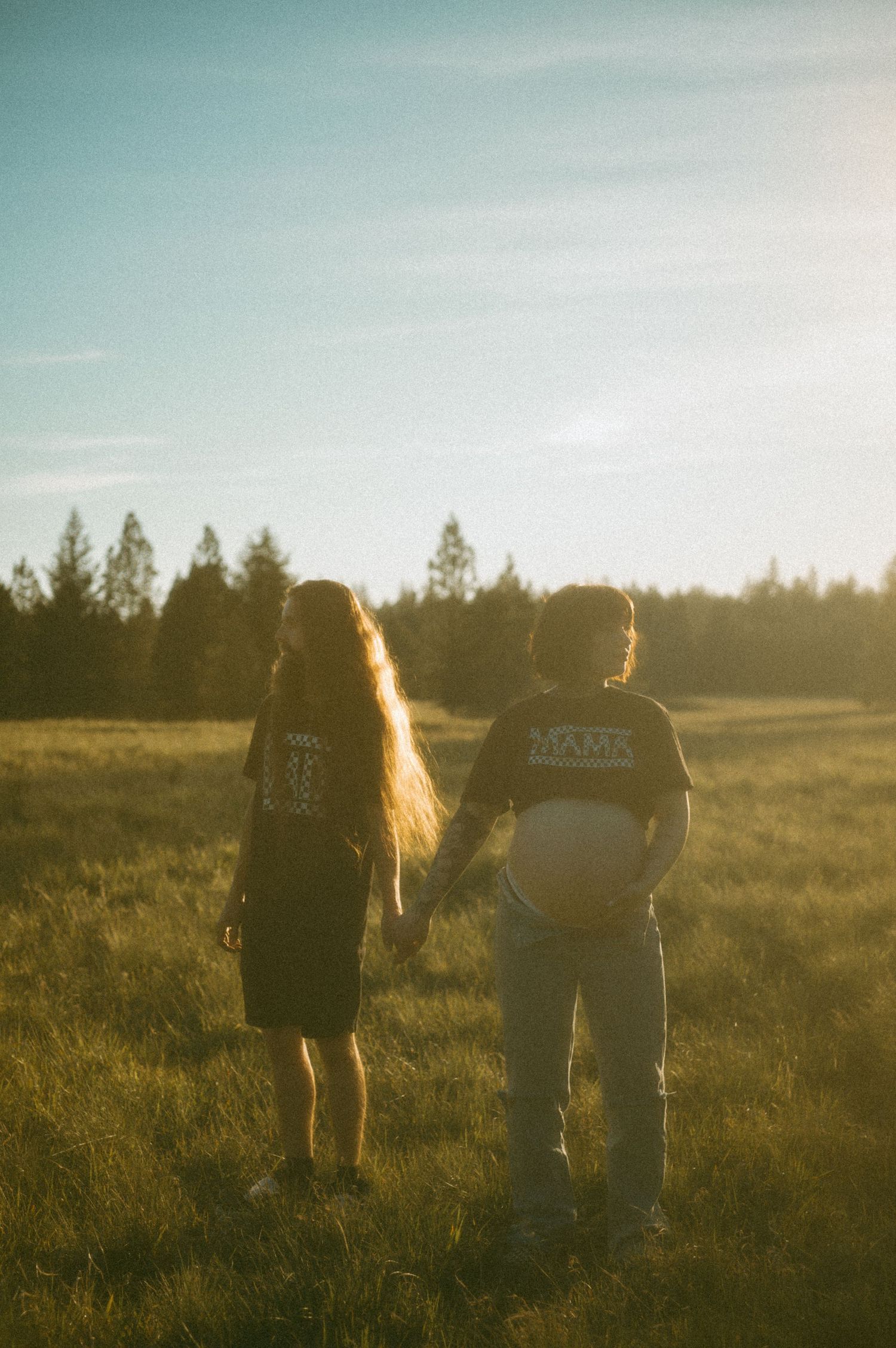 Two people stand together in a sunlit meadow at golden hour with evergreen trees silhouetted in the background.
