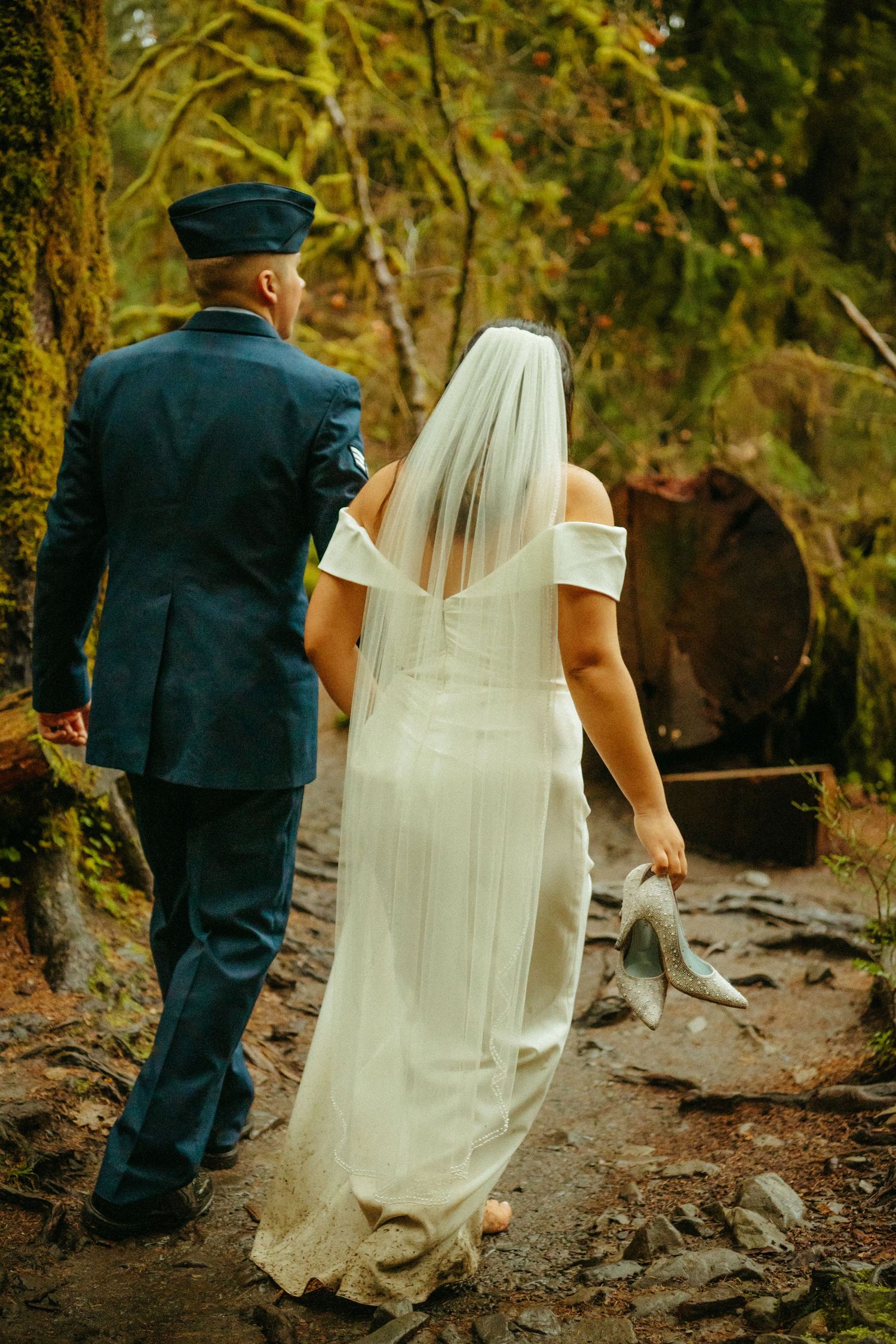 A bride in a white dress and military groom in dress uniform walk together on a wooded path during their wedding.