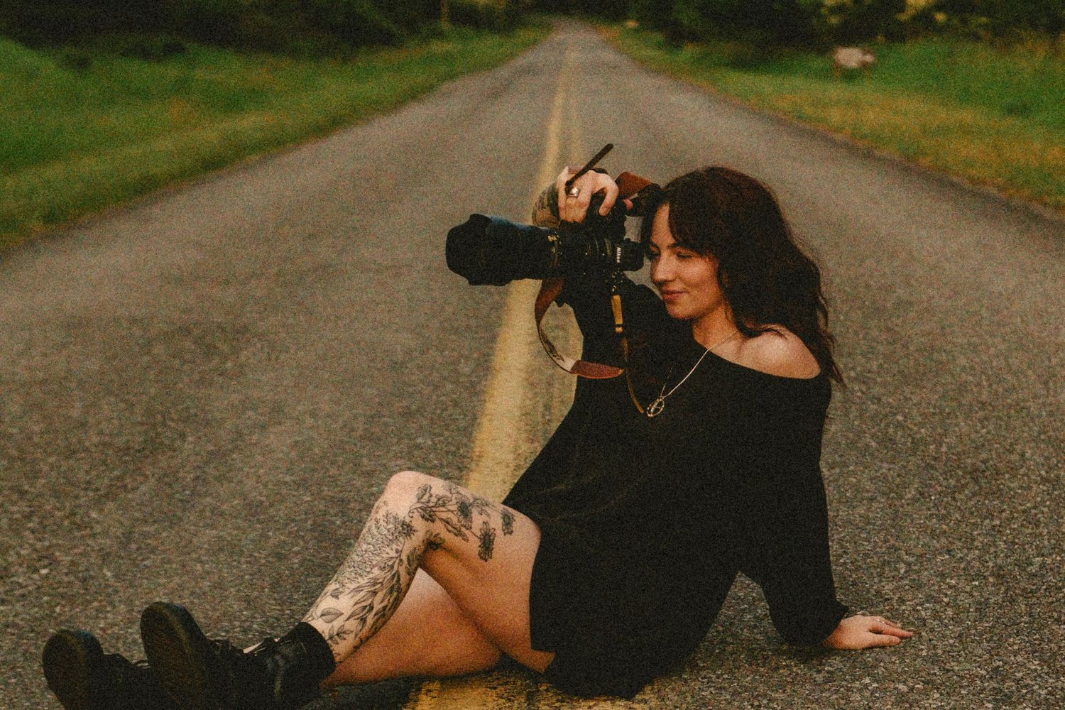 A photographer in black clothing lies on a rural road while taking pictures with a professional camera.