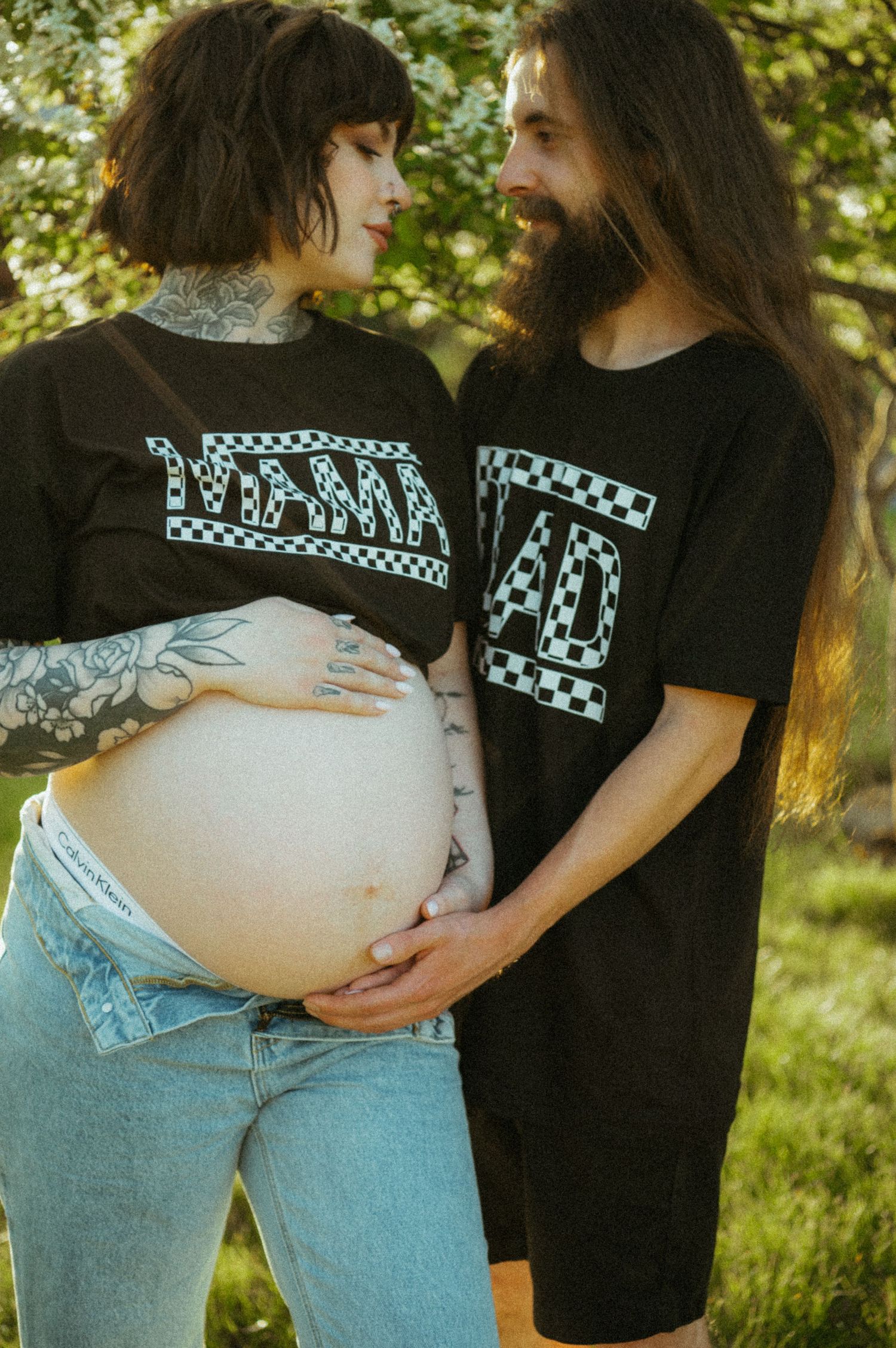 Two people in matching graphic t-shirts stand together outdoors, one with a visible pregnant belly.