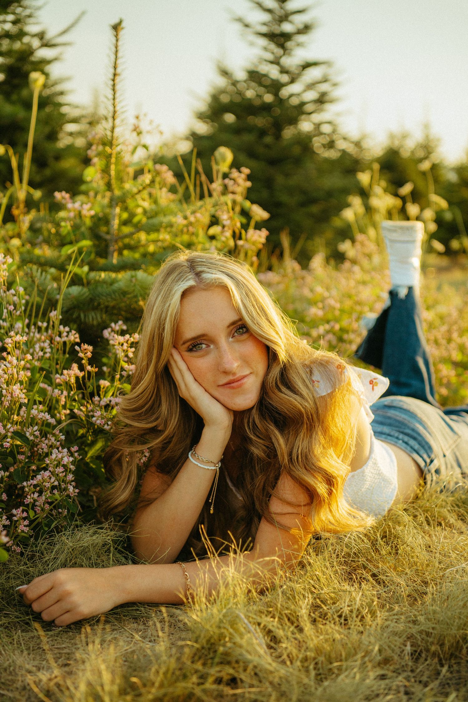 A person lying in a sunny meadow at golden hour surrounded by wildflowers and evergreen trees in the background.