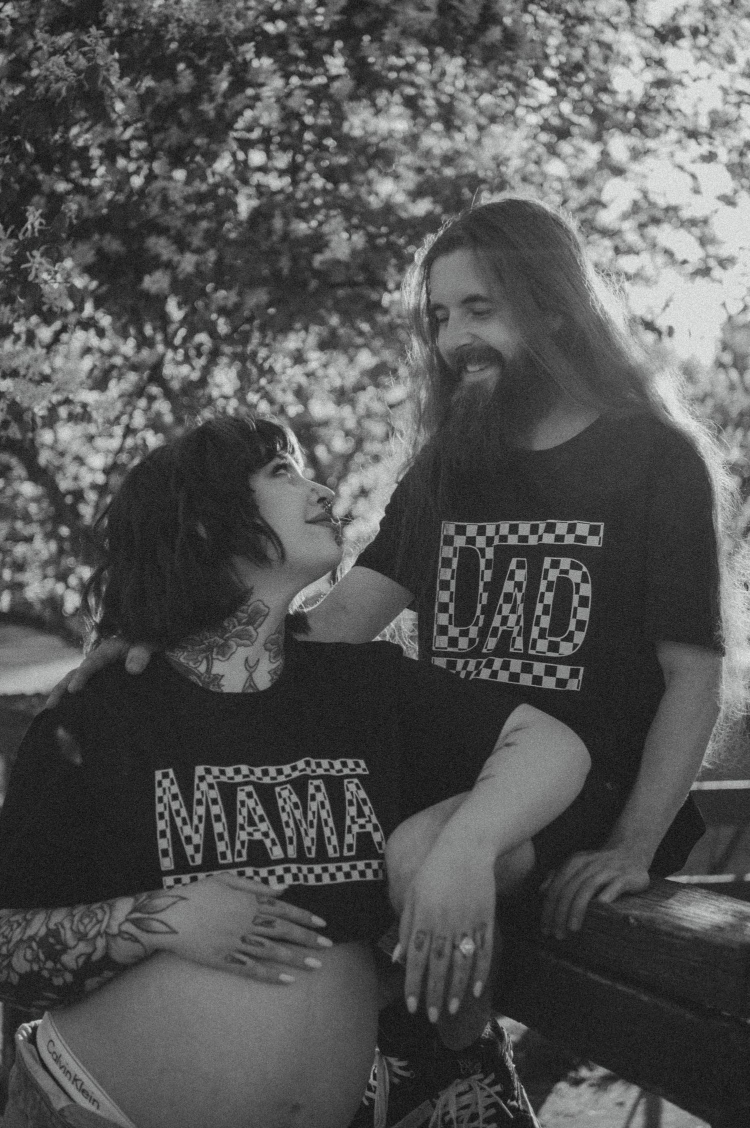 A black and white photograph shows a couple wearing matching graphic t-shirts sharing an intimate moment outdoors.
