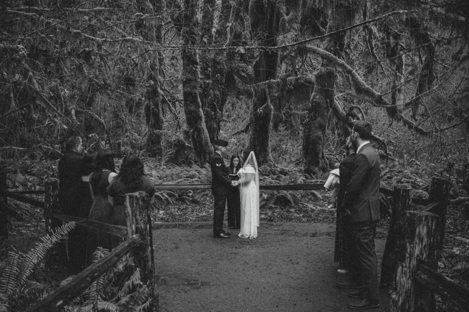 A black and white photograph of an intimate forest wedding ceremony surrounded by tall trees and stone walls.