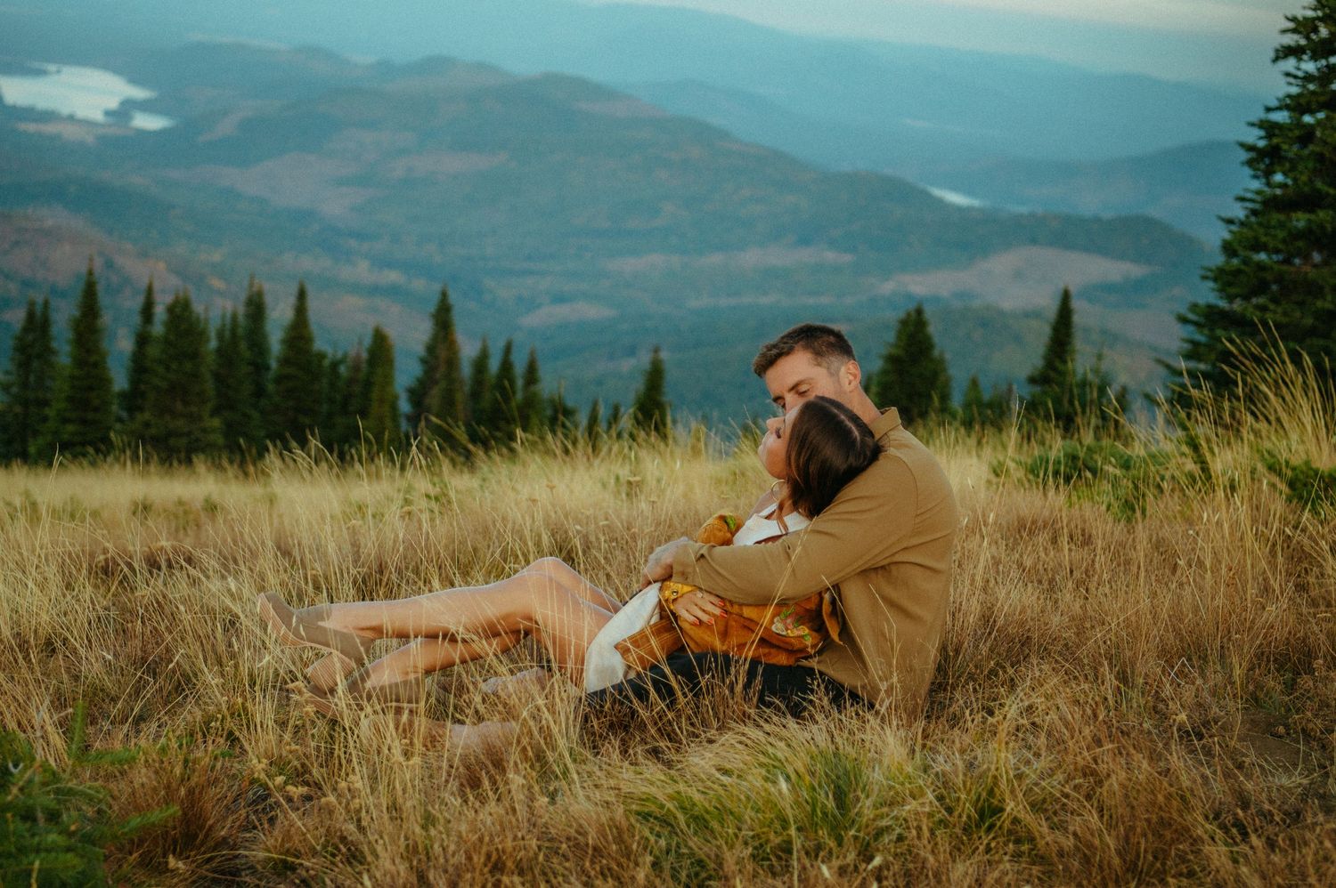 Scenic mountain view with someone in brown coat sitting in tall grass meadow at sunset.