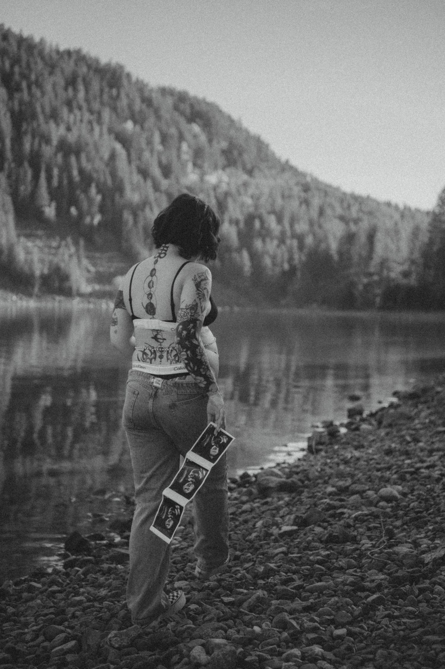 Black and white photo of a person in denim and tank top standing by a serene lake surrounded by forested hills.