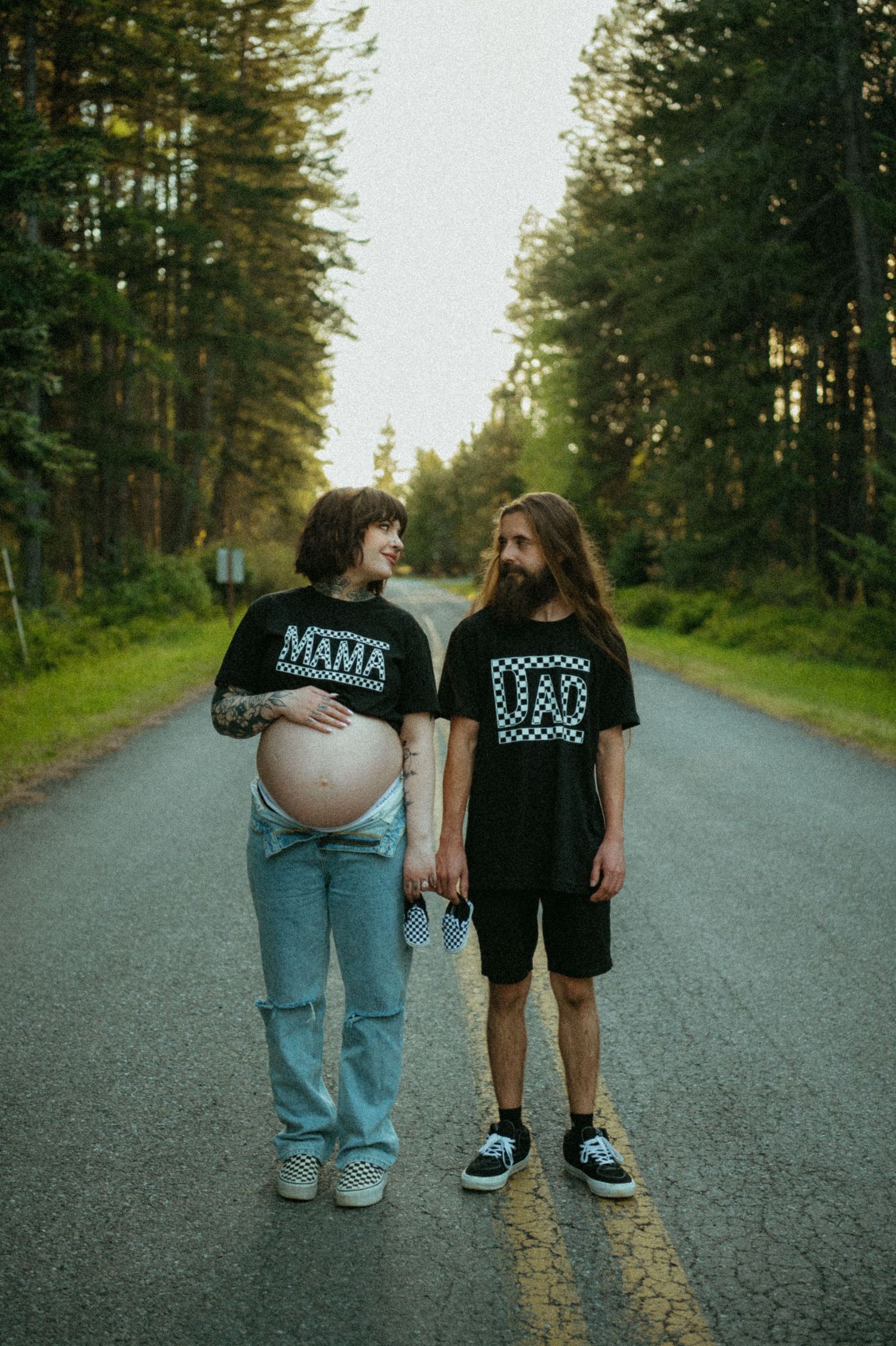 A couple stands together on a forested road wearing matching Mama and Dad t-shirts, expecting a baby.