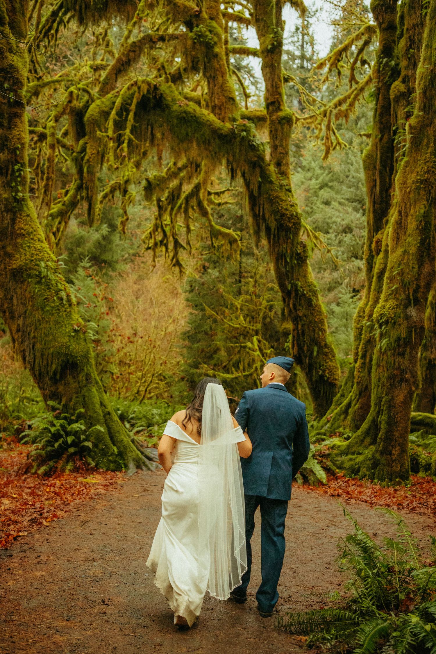 A couple in wedding attire walks down a moss-covered forest path surrounded by ancient trees in the Pacific Northwest.