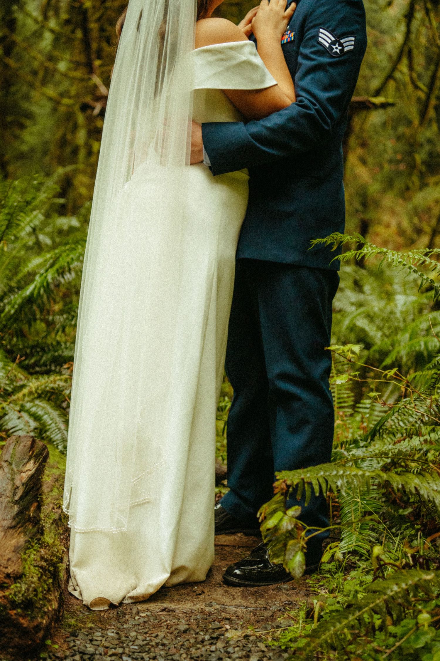 Romantic wedding photo showing couple embracing in a lush forest setting with flowing white veil and navy uniform.
