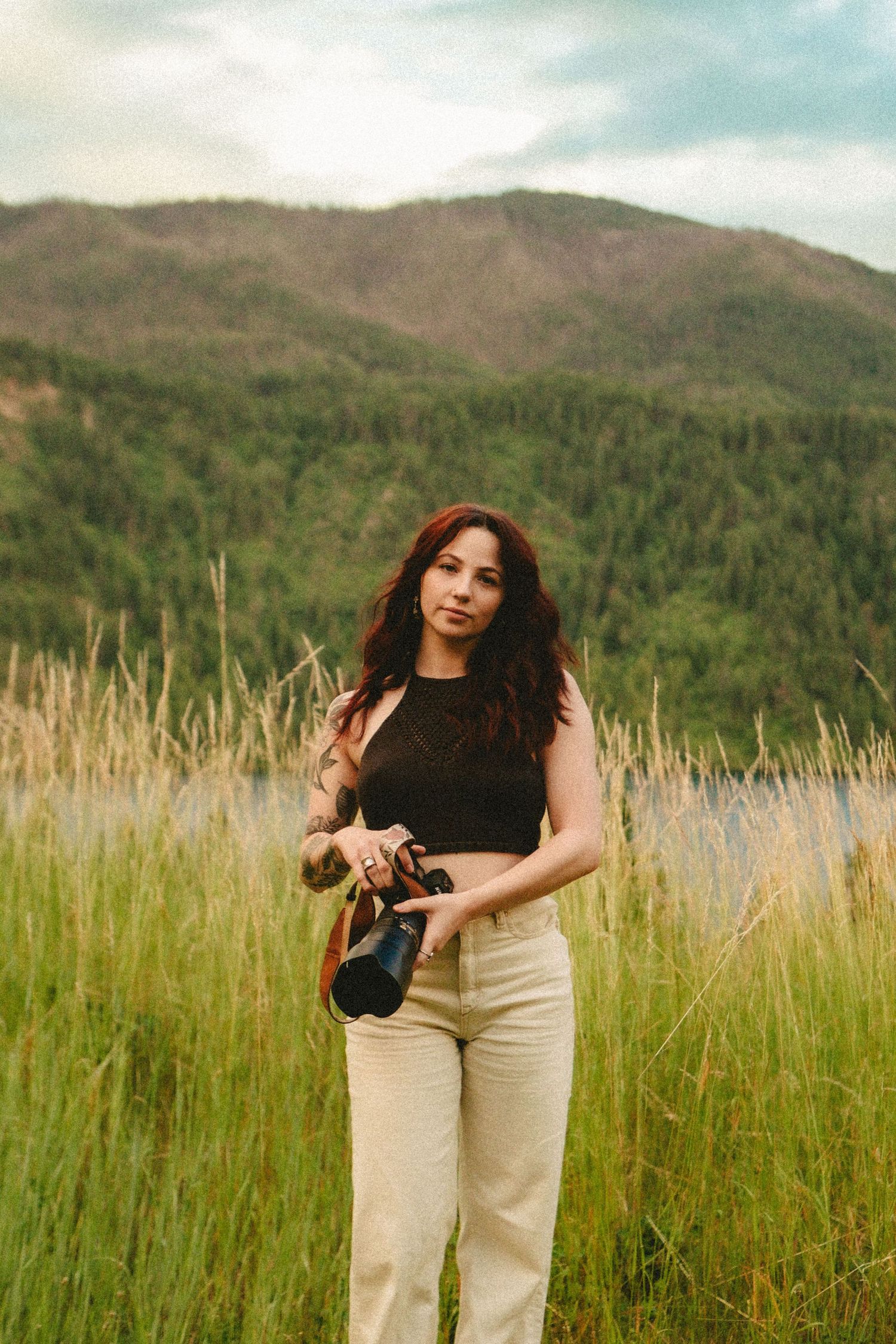 Person in a black crop top and light pants stands in tall grass with mountains and forest in the background.