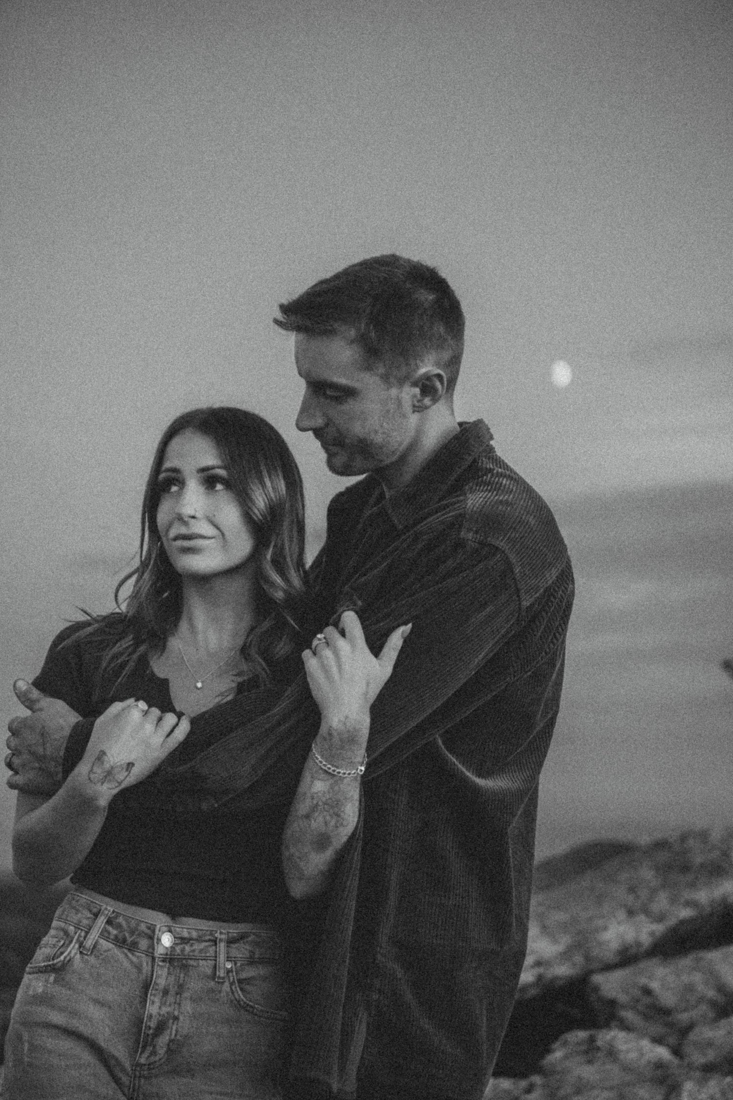 A romantic black and white photo of a couple embracing on a coastal cliff at dusk with the moon visible in the background.