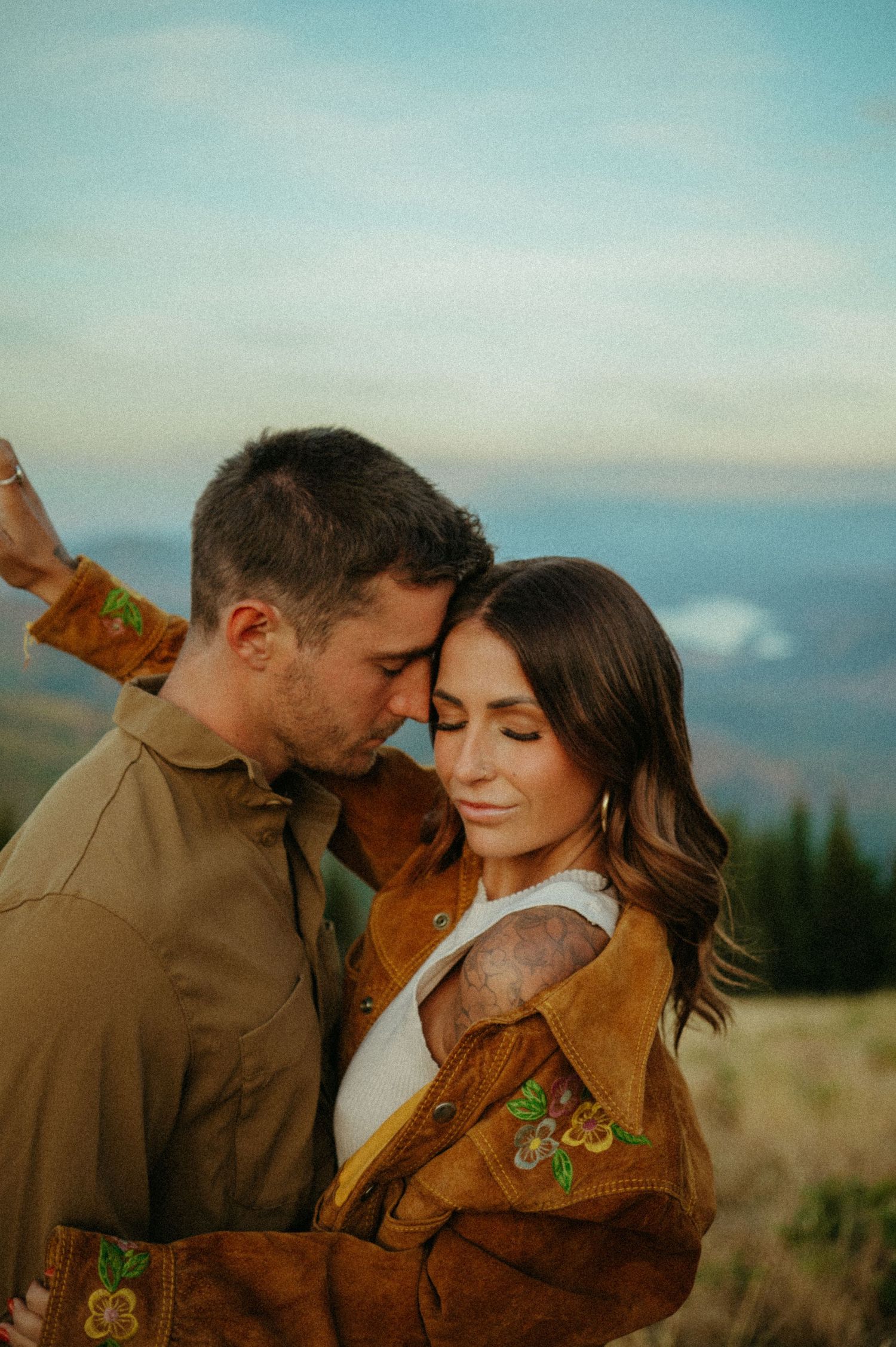 Romantic couple in matching brown jackets embrace on mountainside against blue sky and misty landscape.